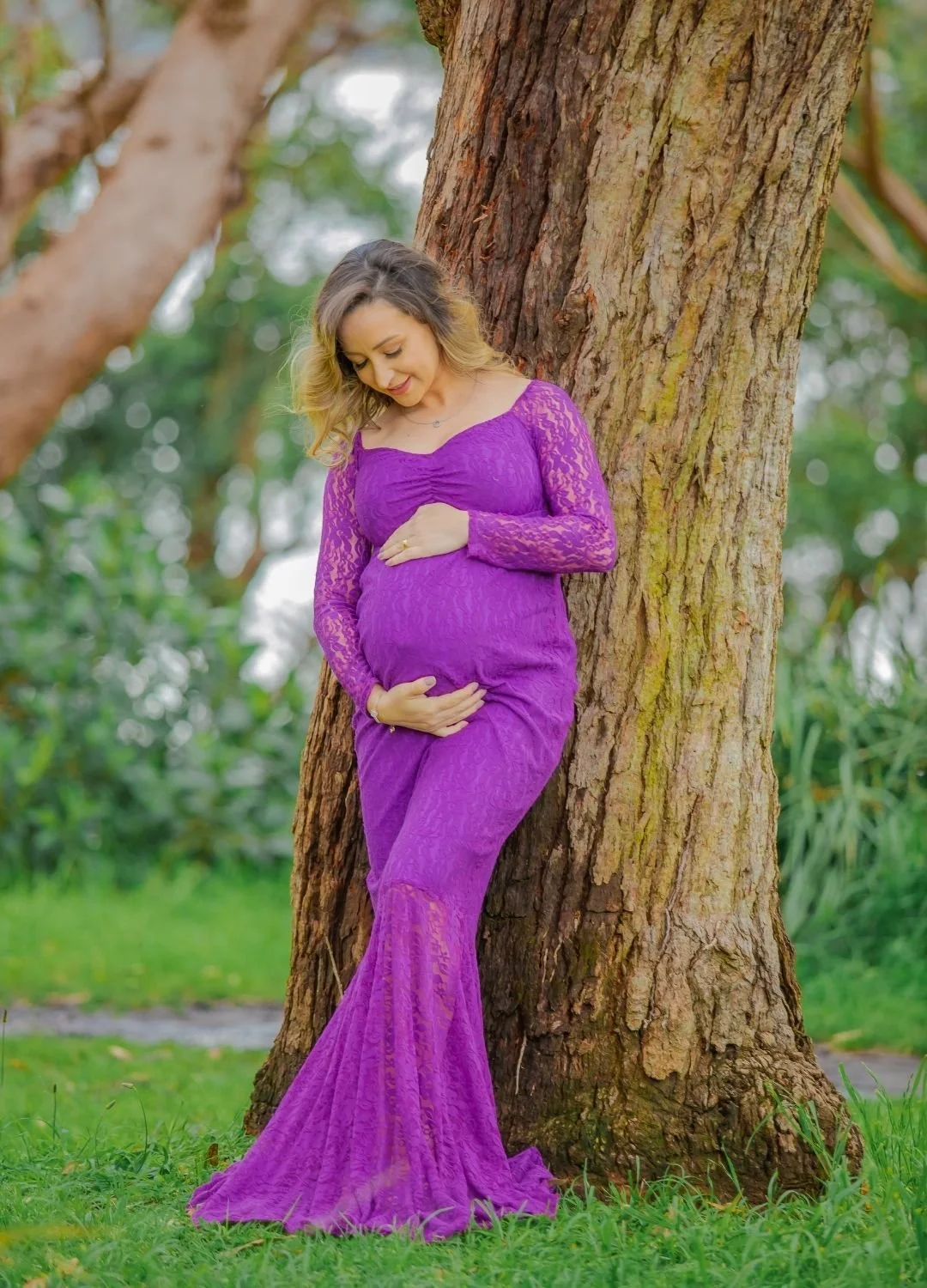 A pregnant woman in a purple lace dress standing outdoors, gently holding her belly with both hands, leaning against a large tree with a background of green foliage.