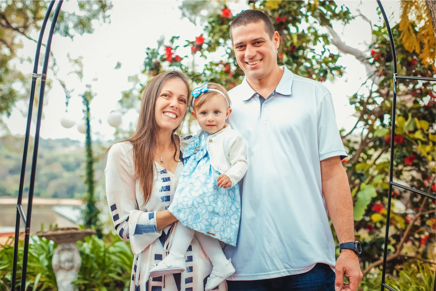 Family of three, mother, father, and young girl, smiling outdoors during daytime with trees and flowers in the background.