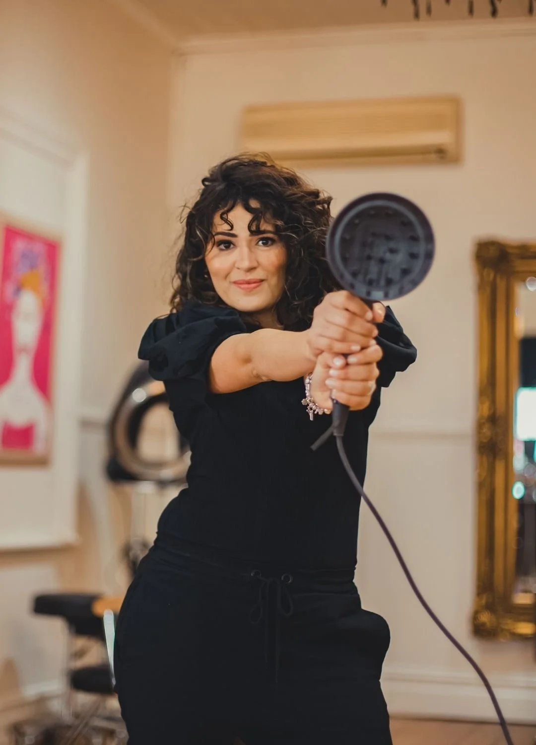 A woman with curly dark hair smiling and holding a hairdryer towards the camera, wearing a black outfit in an indoor setting with a mirror and artwork in the background.