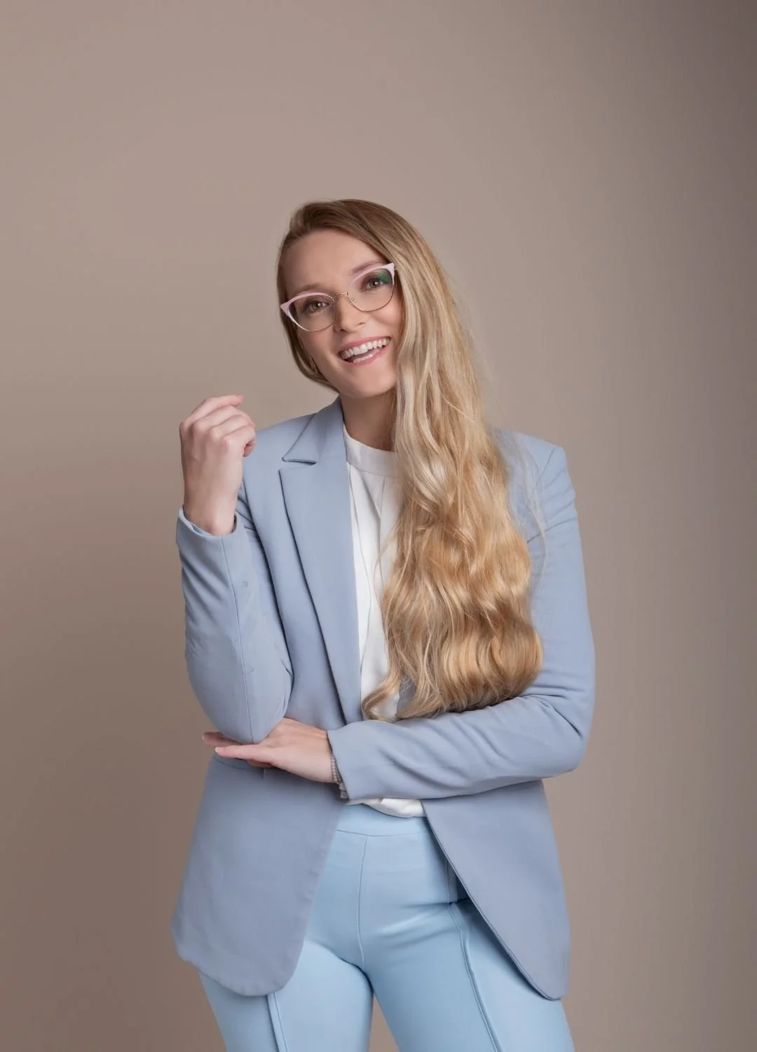 A woman with long blonde hair wearing glasses, a light blue suit, and a white blouse, smiling and posing with one hand near her face against a plain beige background.