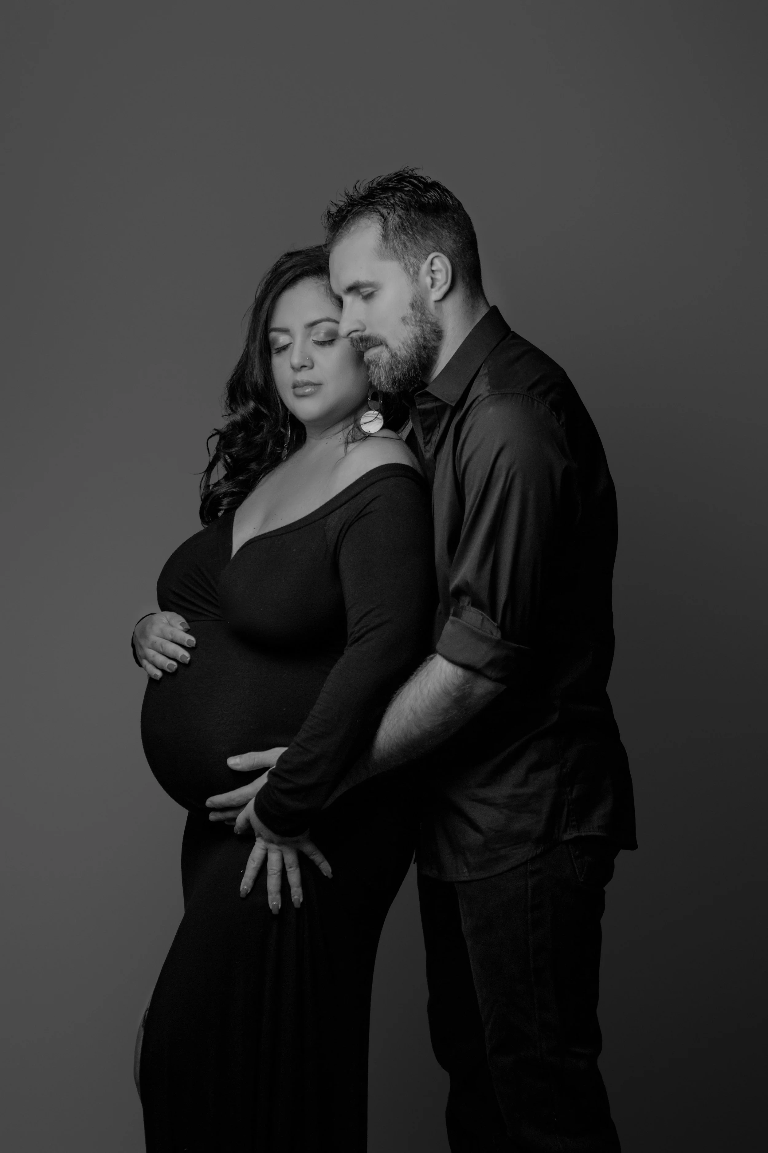 Black and white photo of a pregnant woman and a man standing close with eyes closed, gently touching the woman's belly.