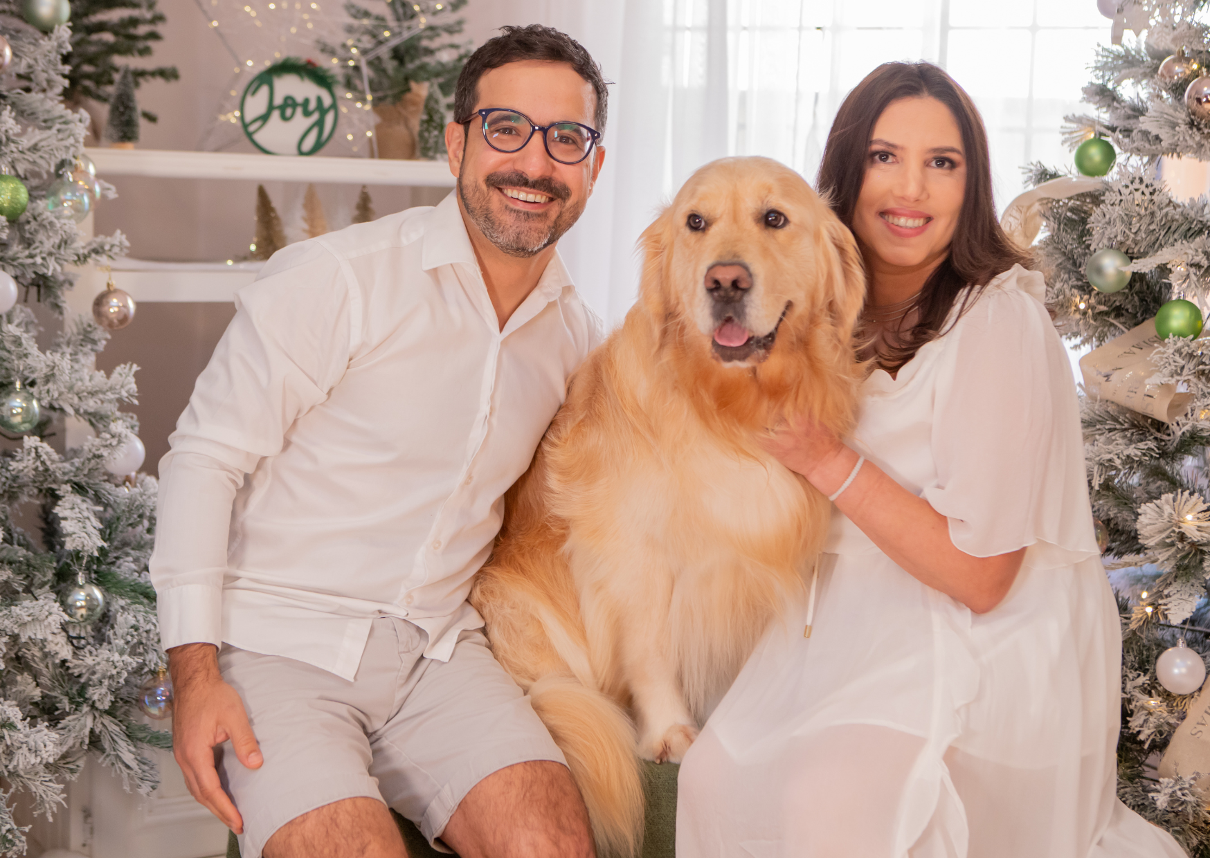 A happy family with a dog, a man, and a woman posing together in front of decorated Christmas trees in a bright, festive room.