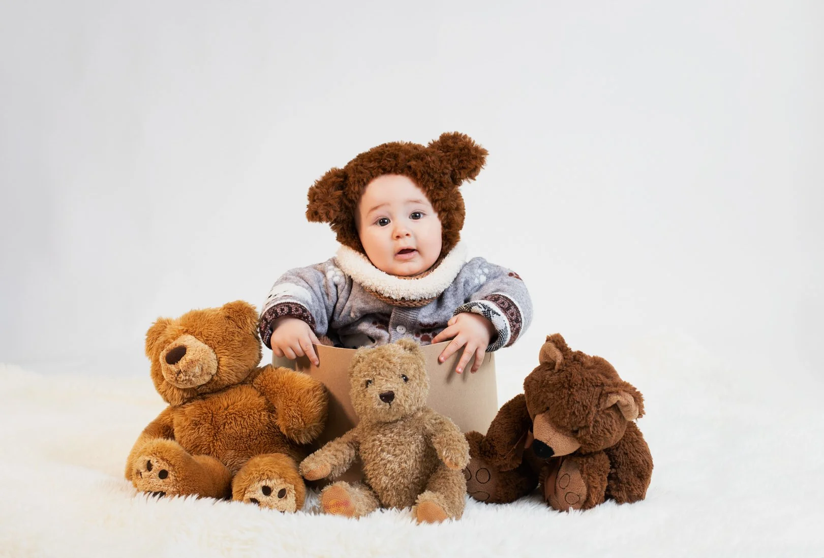 A baby dressed in a cozy sweater with bear ears and a fur collar, sitting among stuffed teddy bears on a soft white surface against a plain white background.