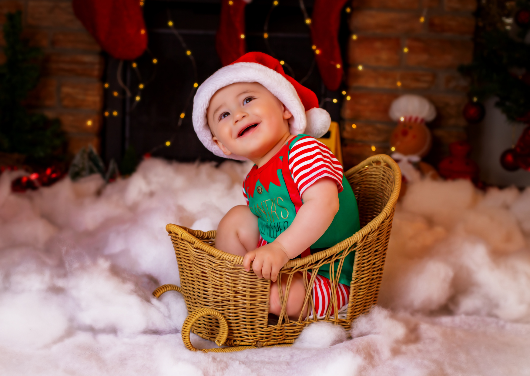 A smiling baby wearing a Santa hat and a festive elf shirt, sitting in a wicker sleigh surrounded by Christmas decorations and fake snow.