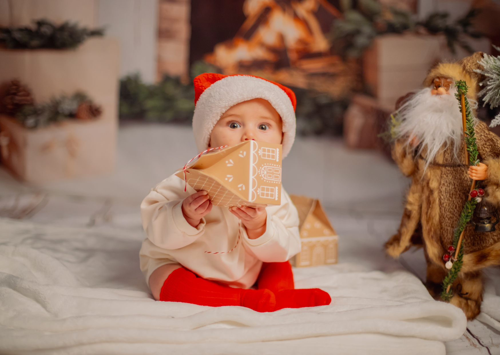 A young child wearing a Santa hat and red socks is sitting on a white blanket, holding and looking surprised at a gift box decorated like a gingerbread house. To the right, there is a Santa doll dressed in a fur coat with a staff, standing near a dec