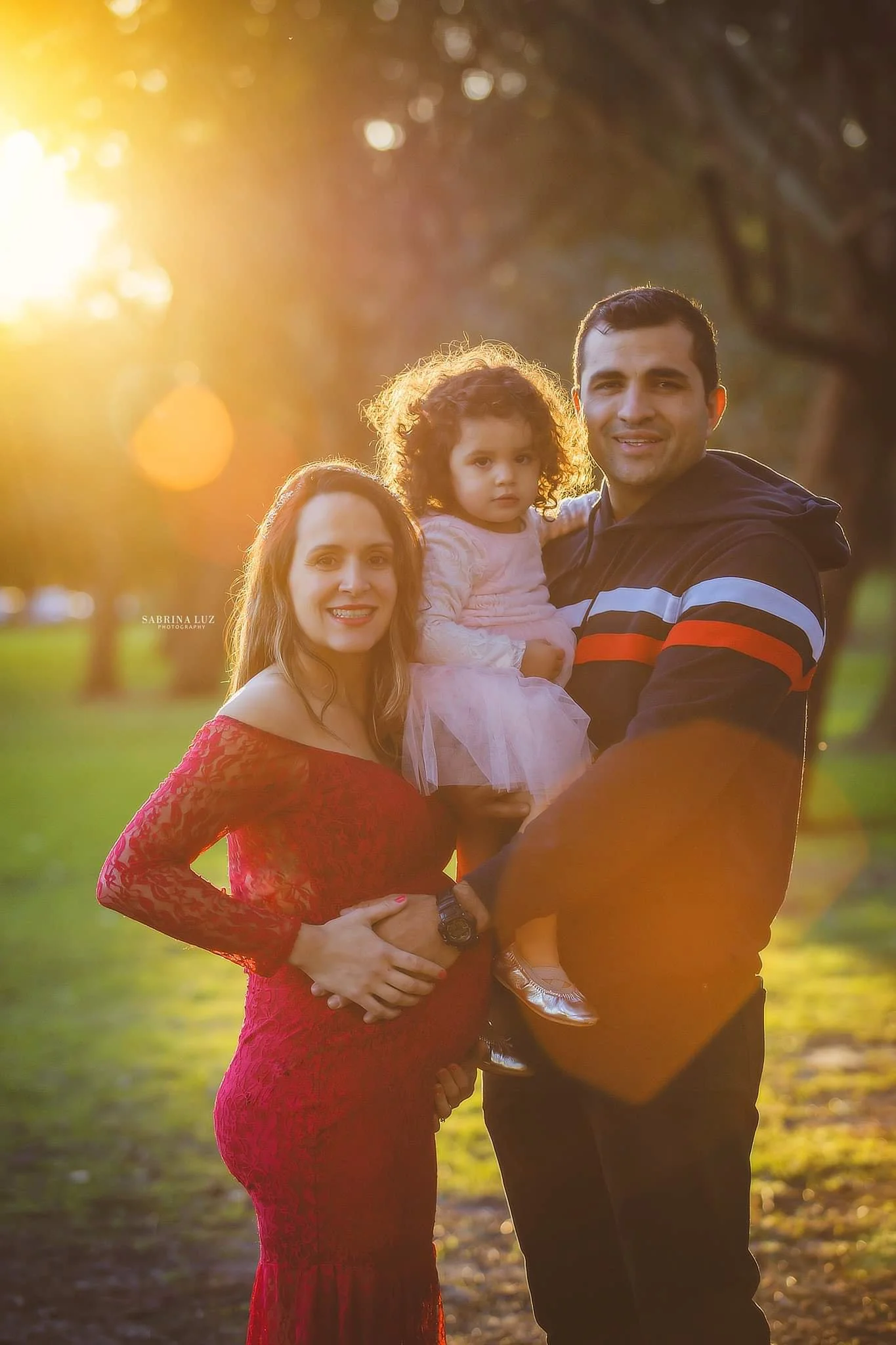 A family of three standing outside during sunset, with a woman in a red dress, a man holding a young girl in a pink dress, surrounded by trees.