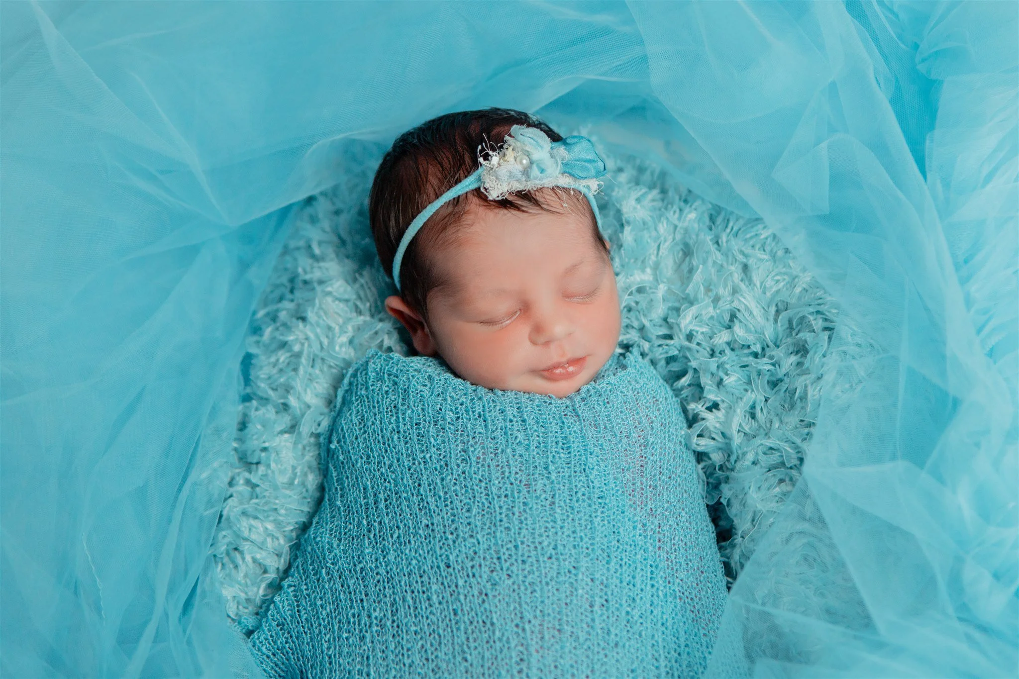 Newborn baby girl sleeping, wrapped in a blue knit blanket, wearing a blue headband with a small floral decoration, surrounded by soft blue tulle fabric.