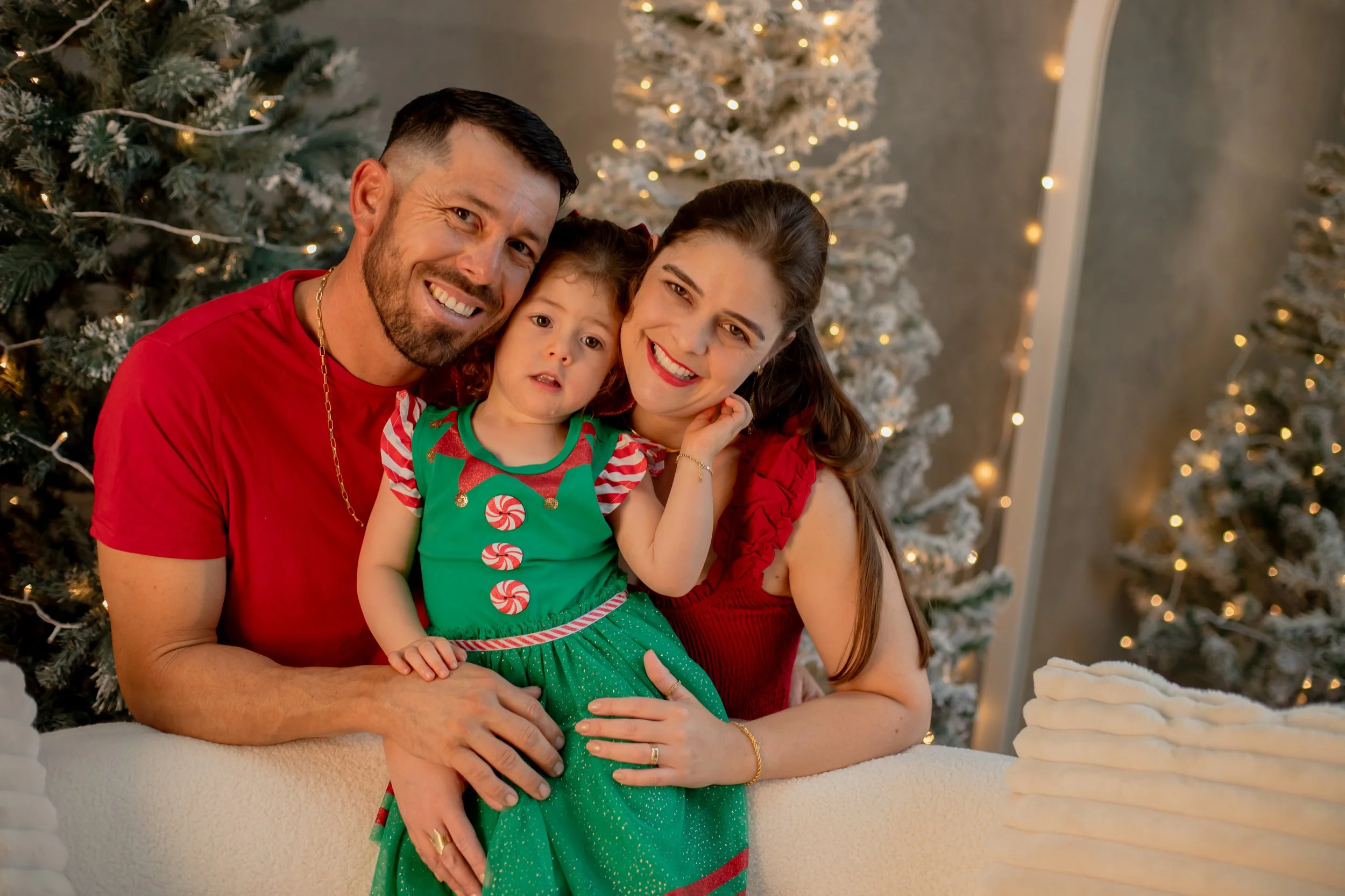 Family of three in Christmas sweaters posing in front of decorated Christmas trees.