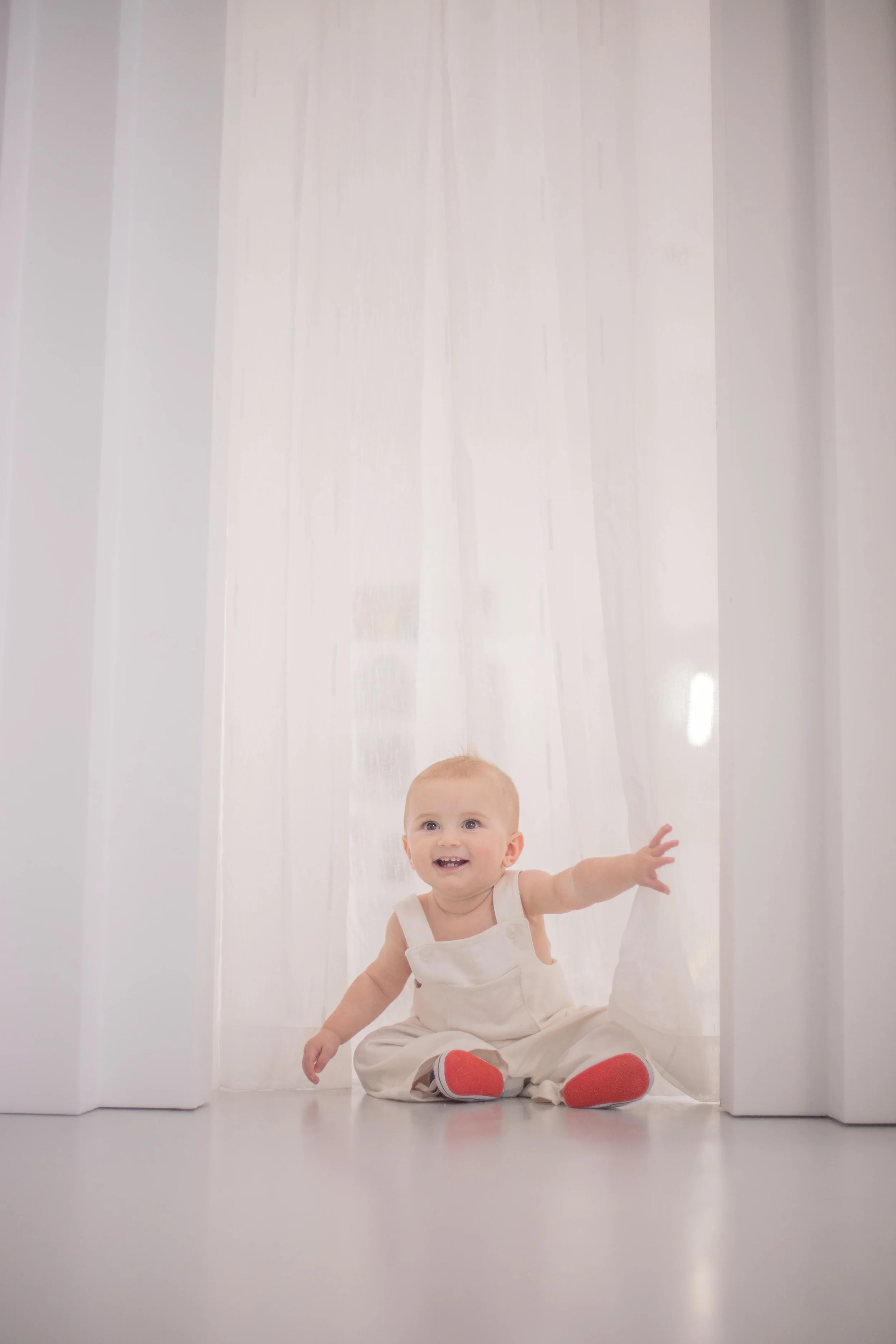 A baby sitting on the floor, reaching out with a joyful expression in front of a sheer curtain.