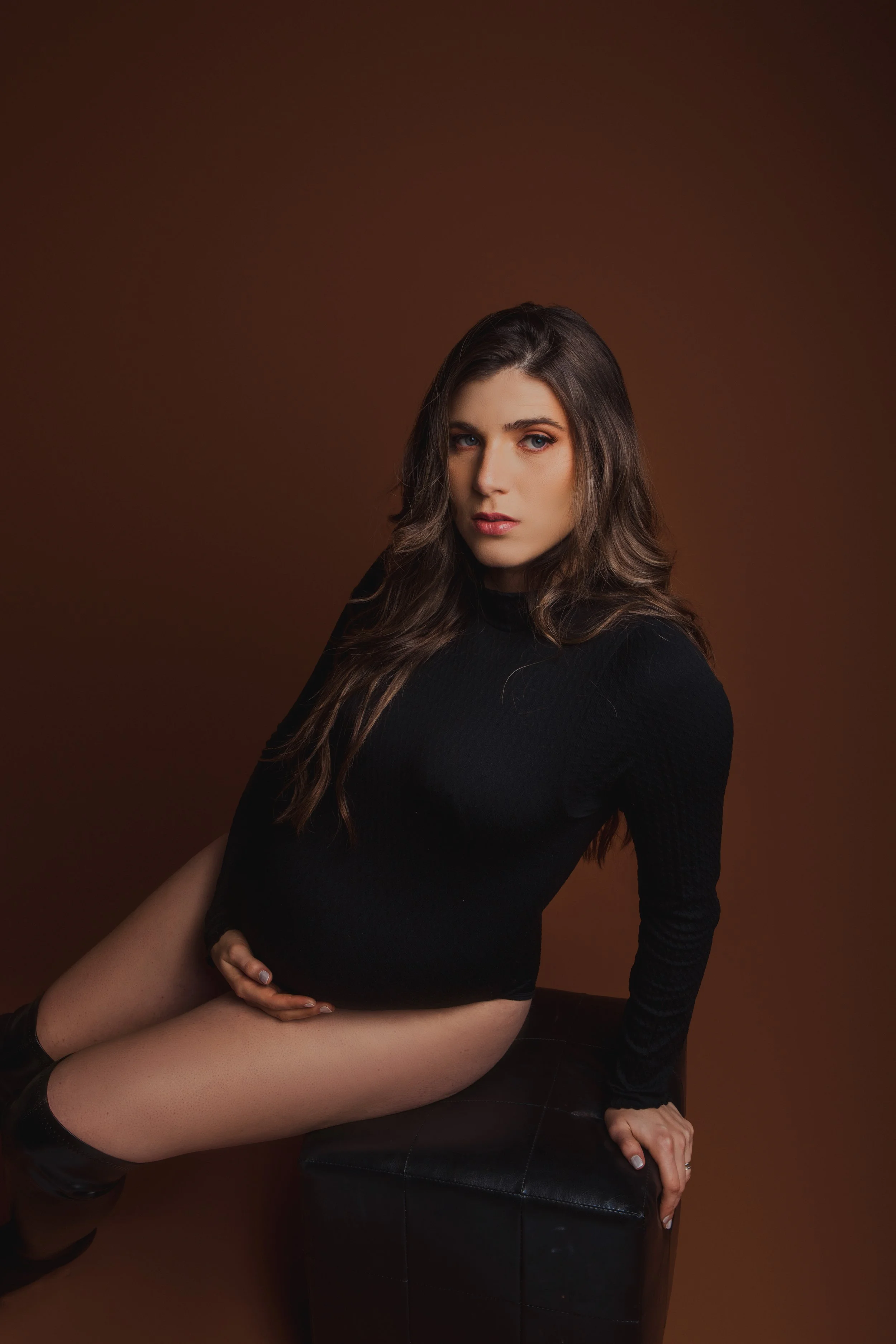 A woman with long, wavy brown hair wearing a black long-sleeve shirt poses sitting on a black leather ottoman against a brown background.