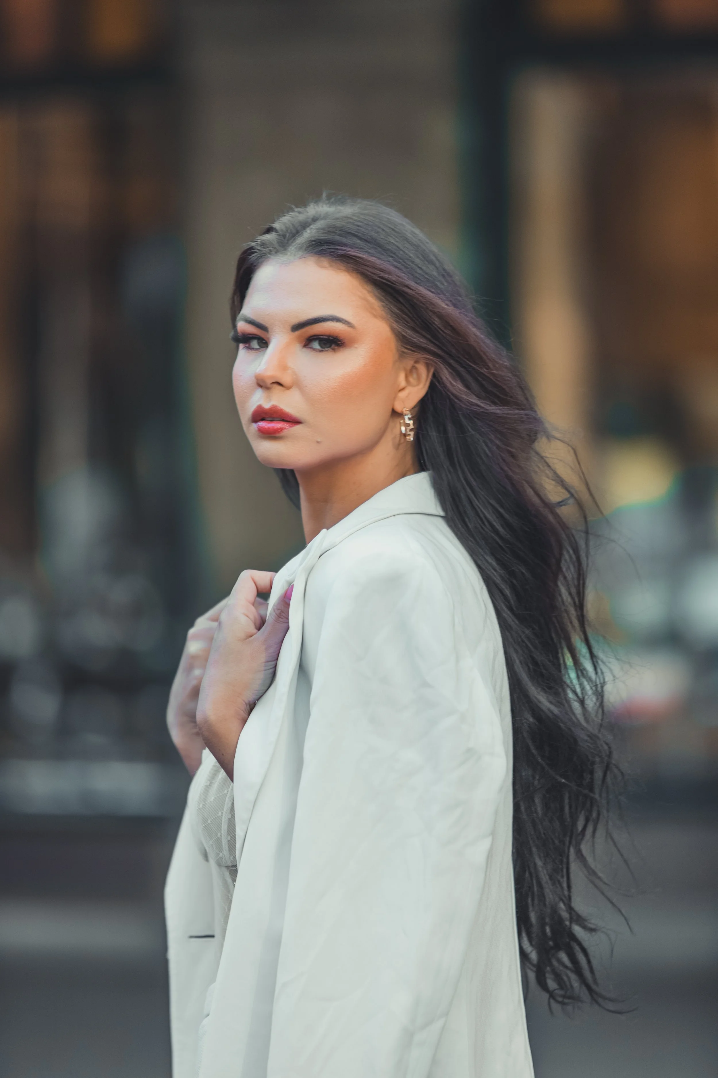 A woman with long dark hair, wearing a white blazer, stands outdoors in an urban setting. She has a confident expression with makeup and earrings, and is looking at the camera.