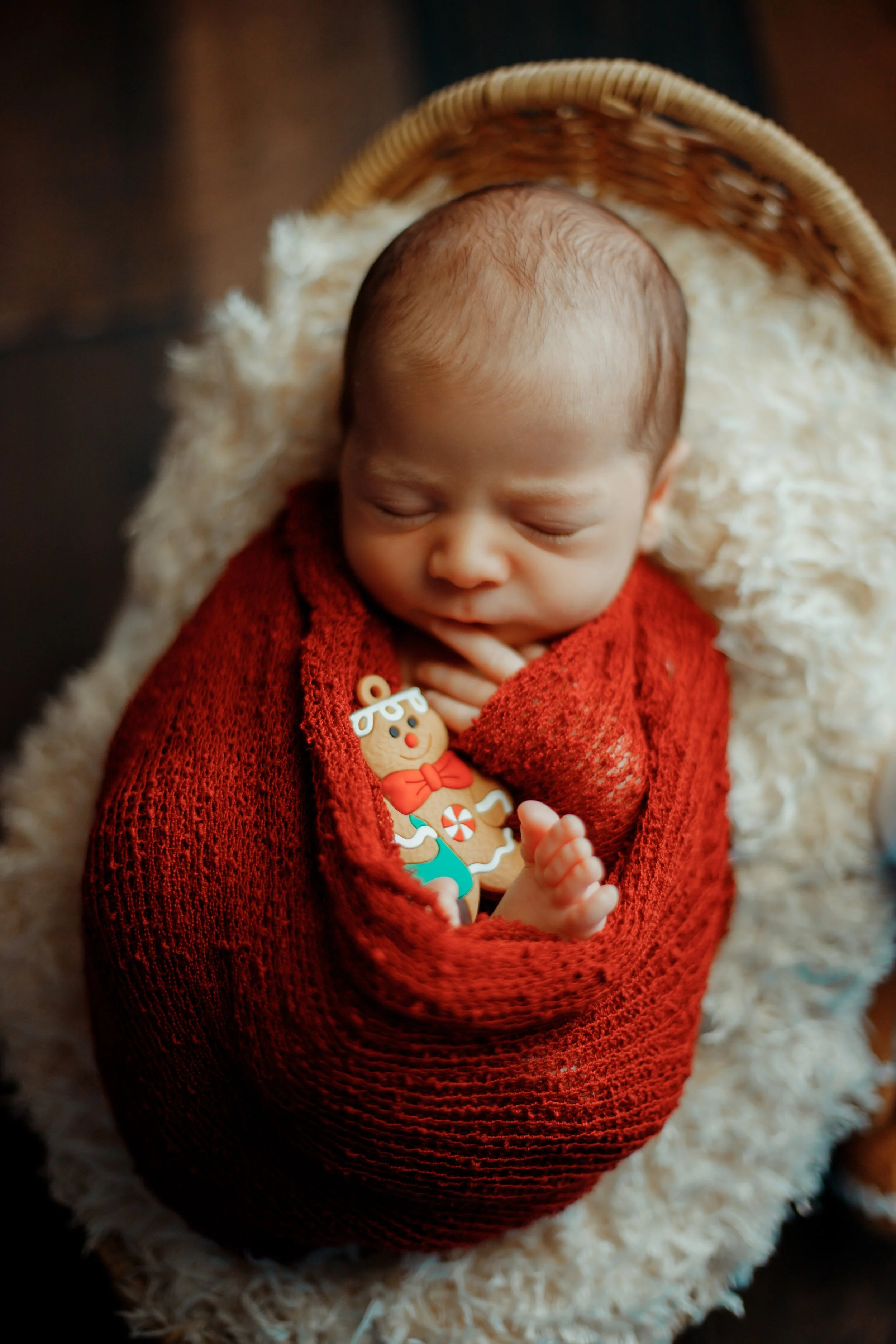 A sleeping baby wrapped in a red knit blanket, lying in a basket with a fluffy cream-colored lining, holding a gingerbread man ornament.