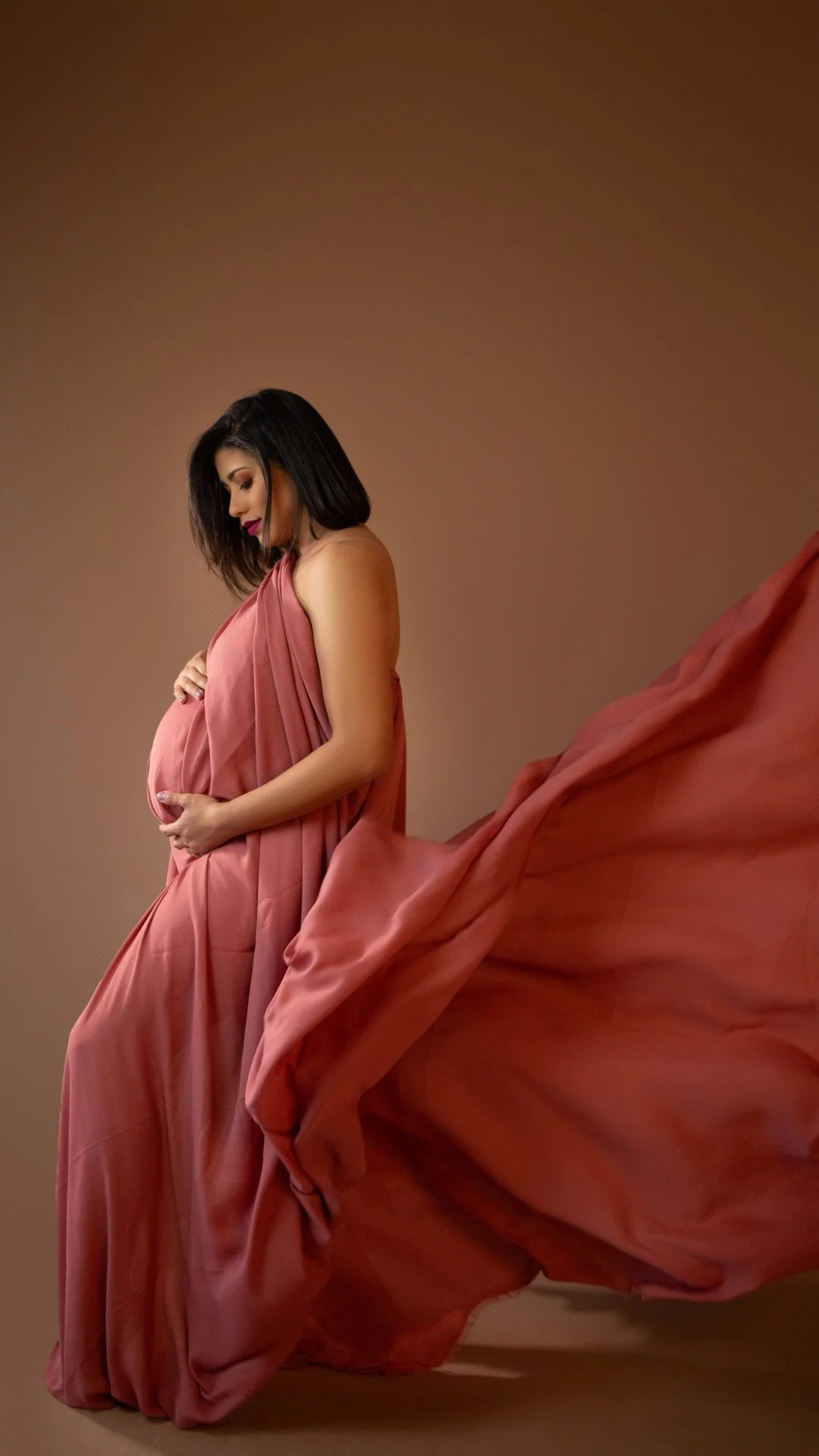 Pregnant woman in a flowing pink dress gently holding her belly with both hands, looking down affectionately.
