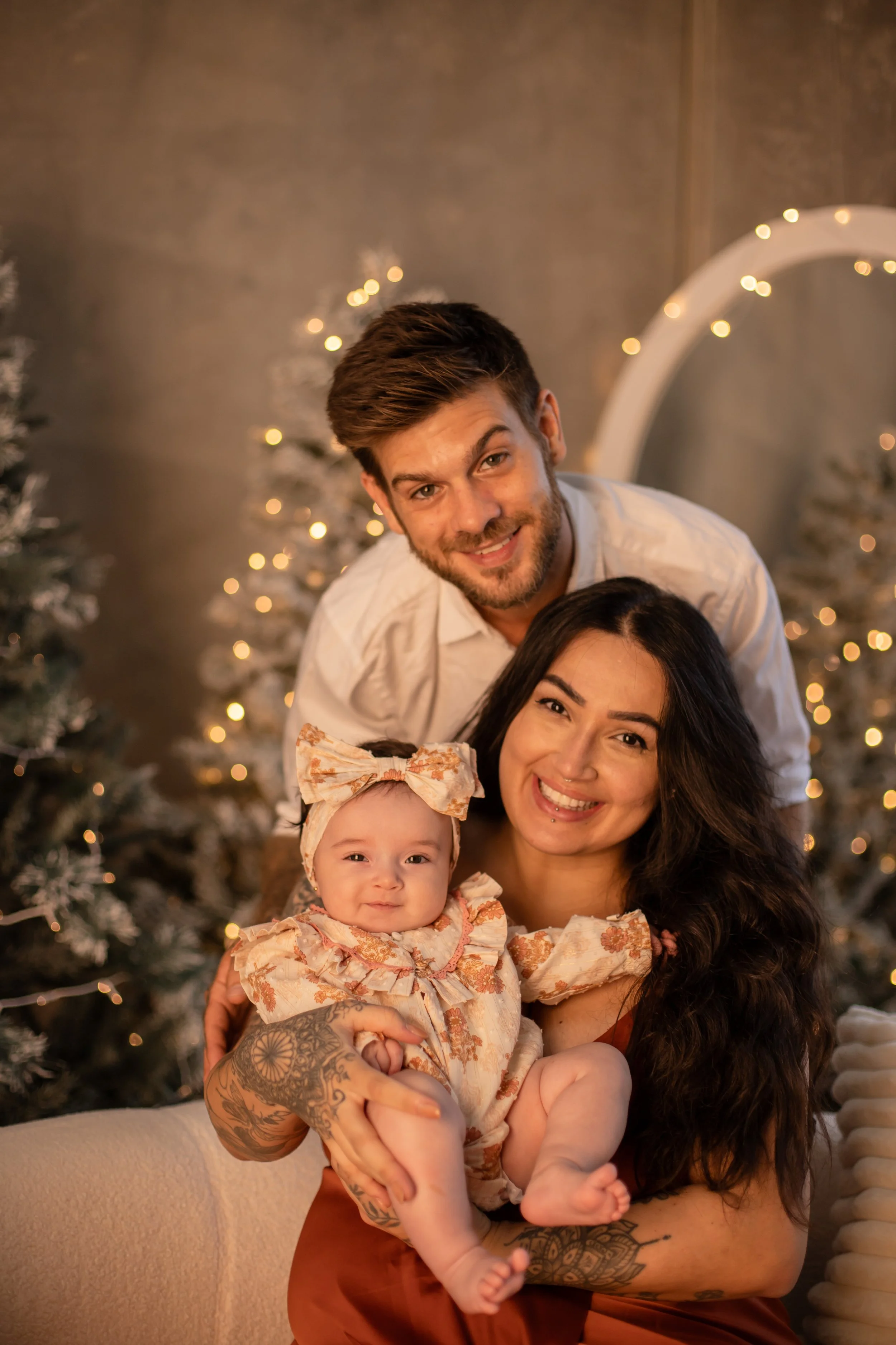 A happy family of three celebrating Christmas, with decorated Christmas trees and string lights in the background.