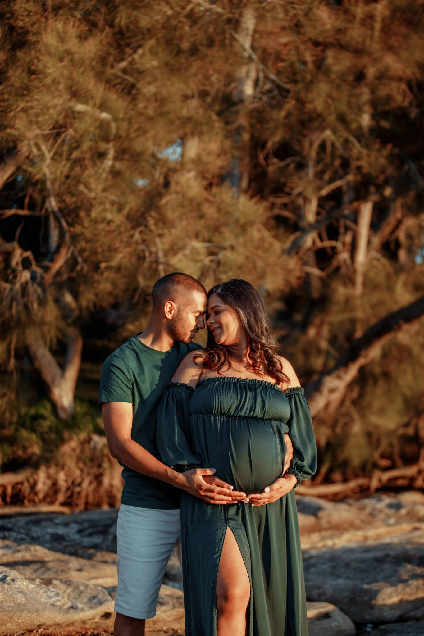 A pregnant woman in a green off-shoulder dress and a man in a green shirt and white shorts embrace on a rocky outdoors setting, their foreheads touching, smiling, with trees in the background.