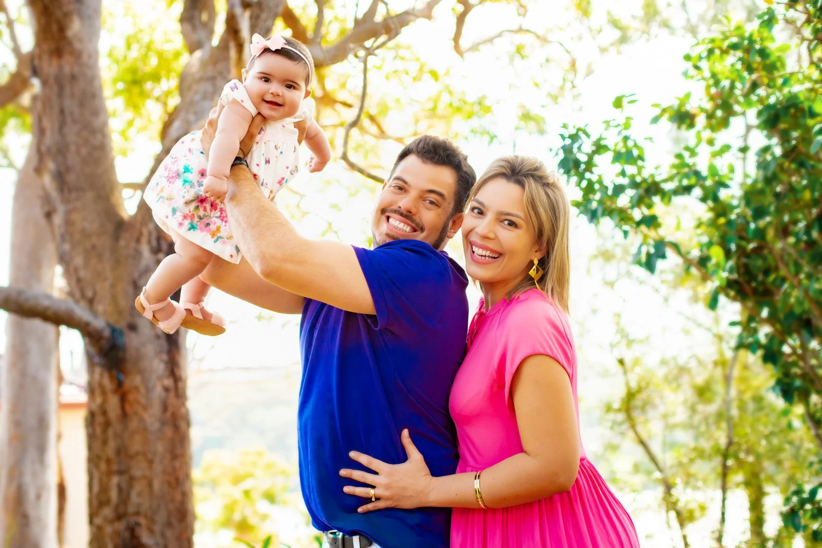 A smiling family of three outdoors, with a man holding a young girl in a floral dress and bow, and a woman wearing a pink dress, all in a green, sunny setting.