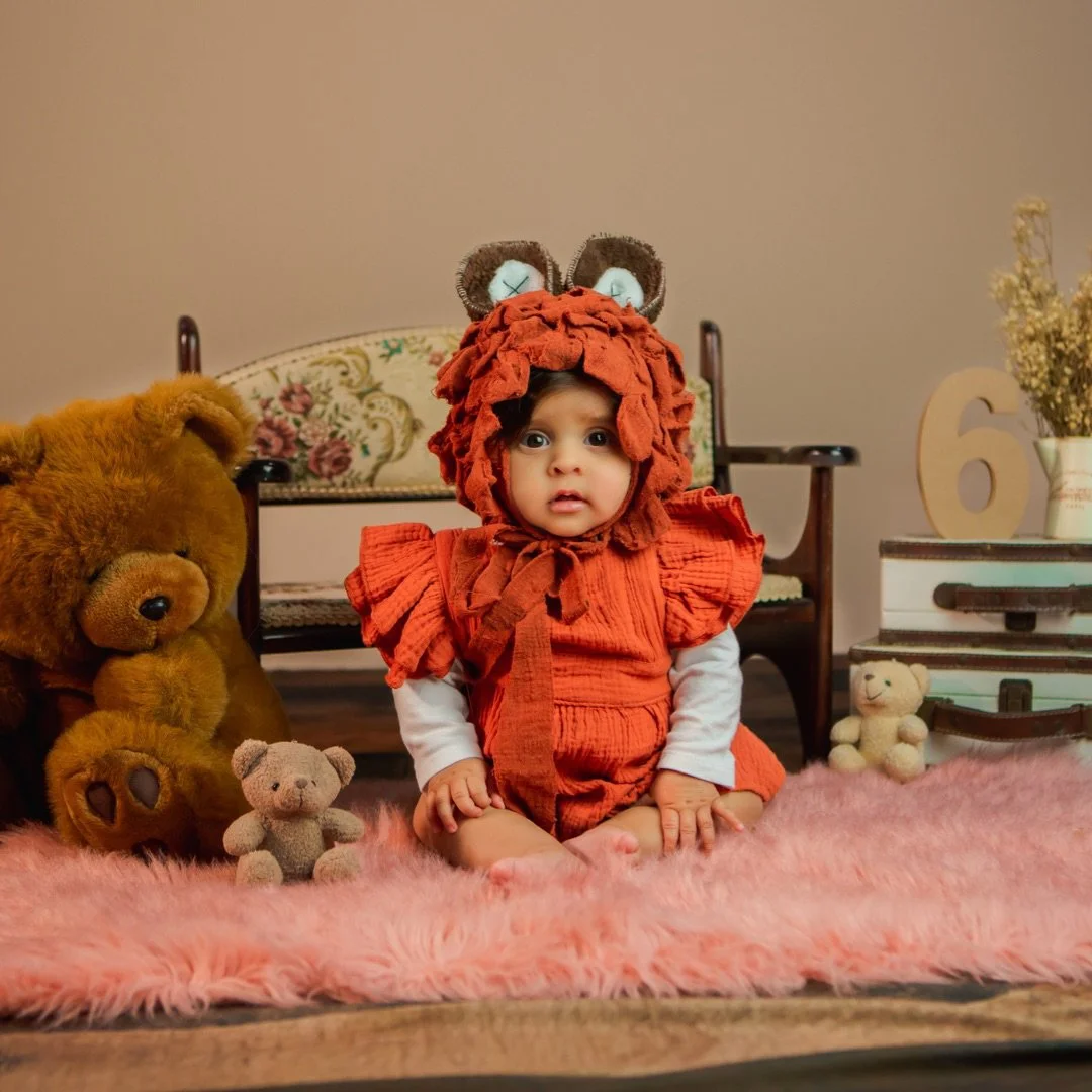 A young child dressed as a lion, sitting on a pink fluffy rug surrounded by teddy bears, with a wooden bench, a flower vase, and stacked boxes in the background.