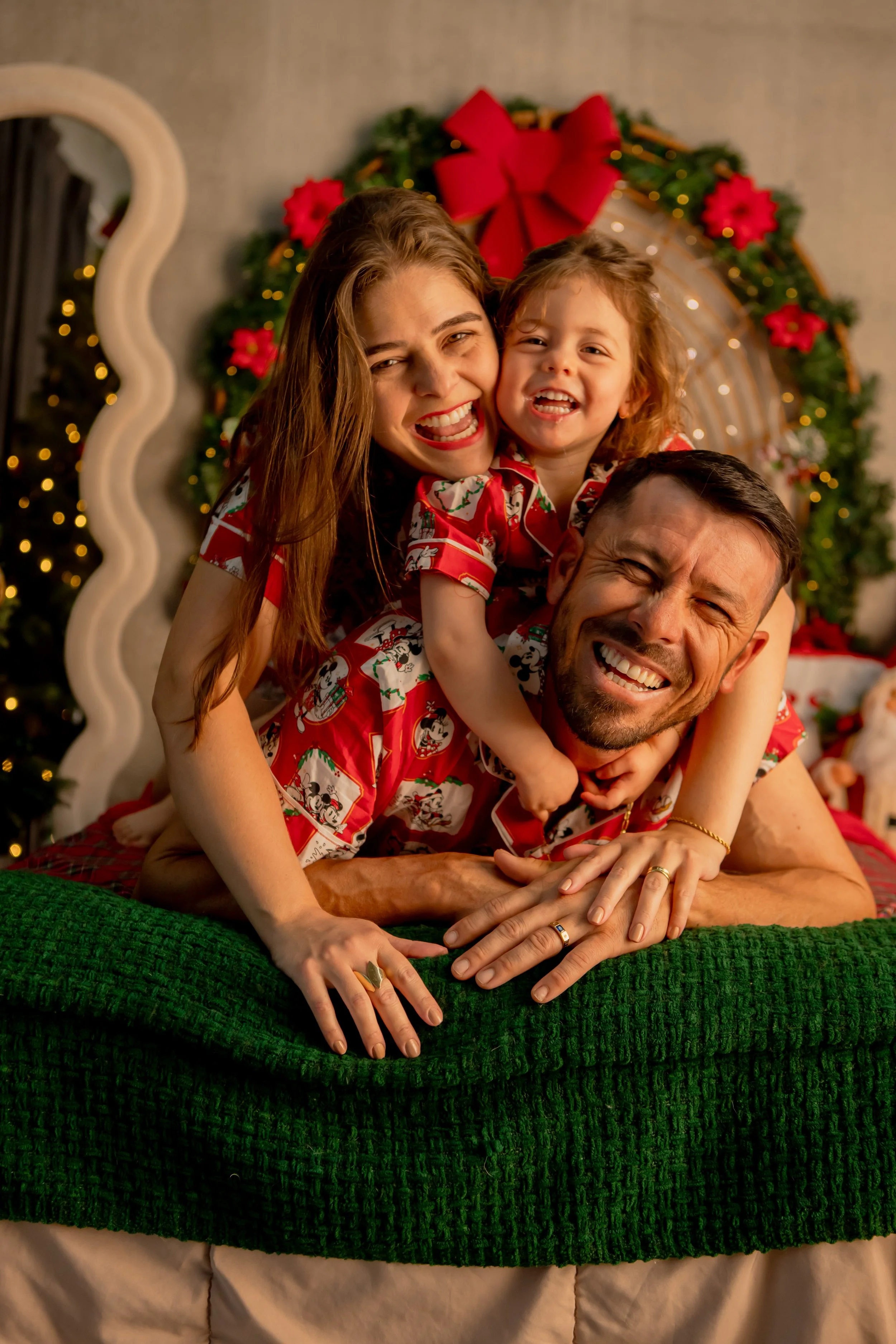 Family of three celebrating Christmas, all wearing matching Christmas pajamas, smiling and playing on a bed with festive holiday decorations in the background.