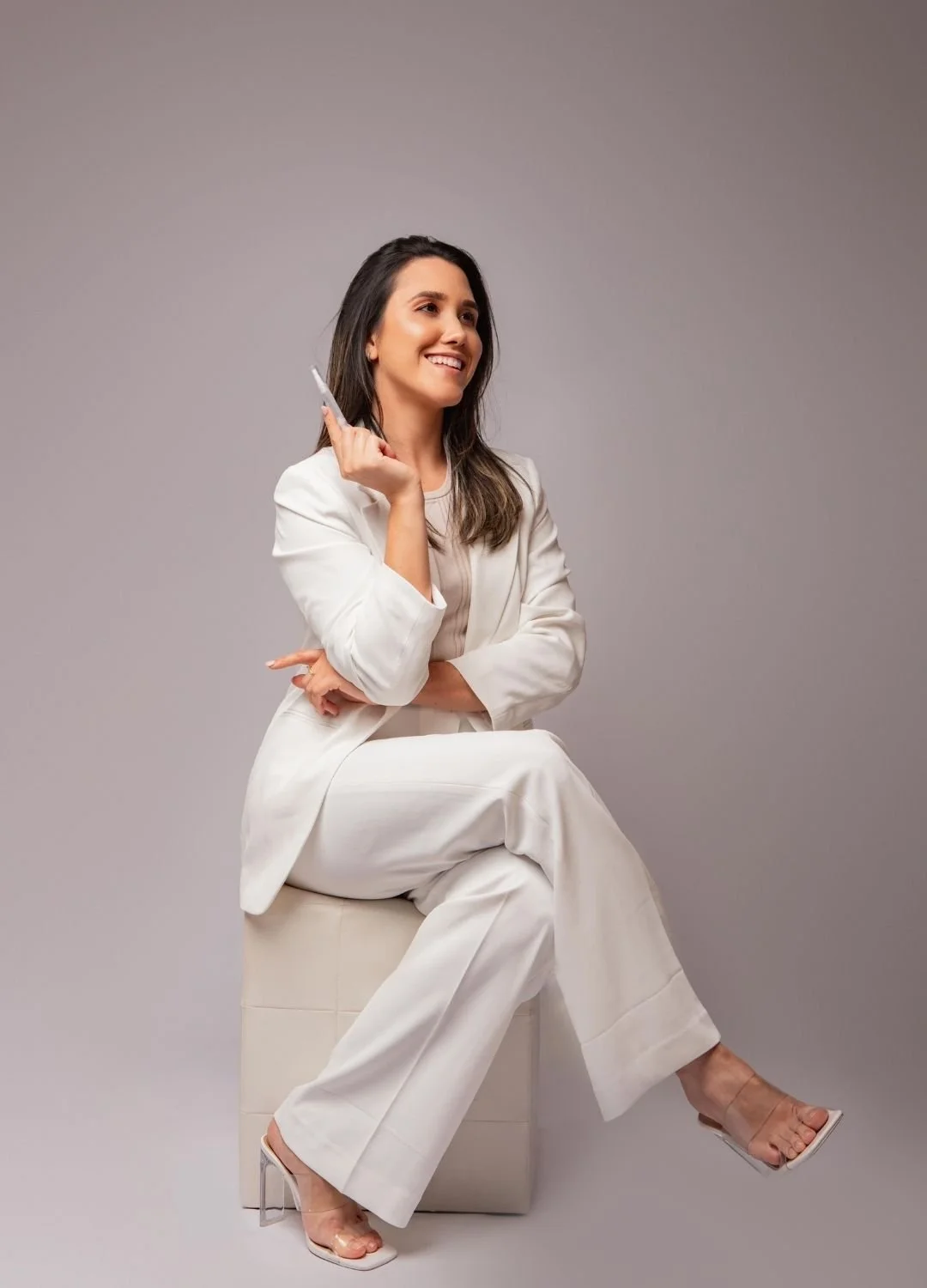 A woman in a white suit sitting on a stool, smiling, holding a pen in her right hand with her arm bent and crossed over her lap, against a plain gray background.