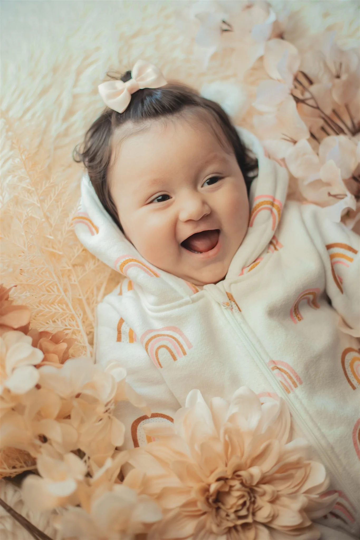 A smiling baby girl with dark hair, wearing a cream-colored hoodie with rainbow patterns, lying on a soft beige surface surrounded by light peach and pink flowers.