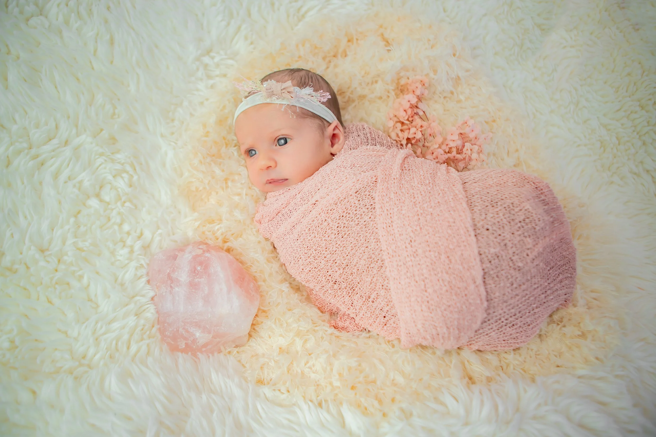 Baby girl lying on a fluffy cream-colored blanket, wearing a pink knitted outfit and a headband with flowers, with a pink salt lamp nearby.