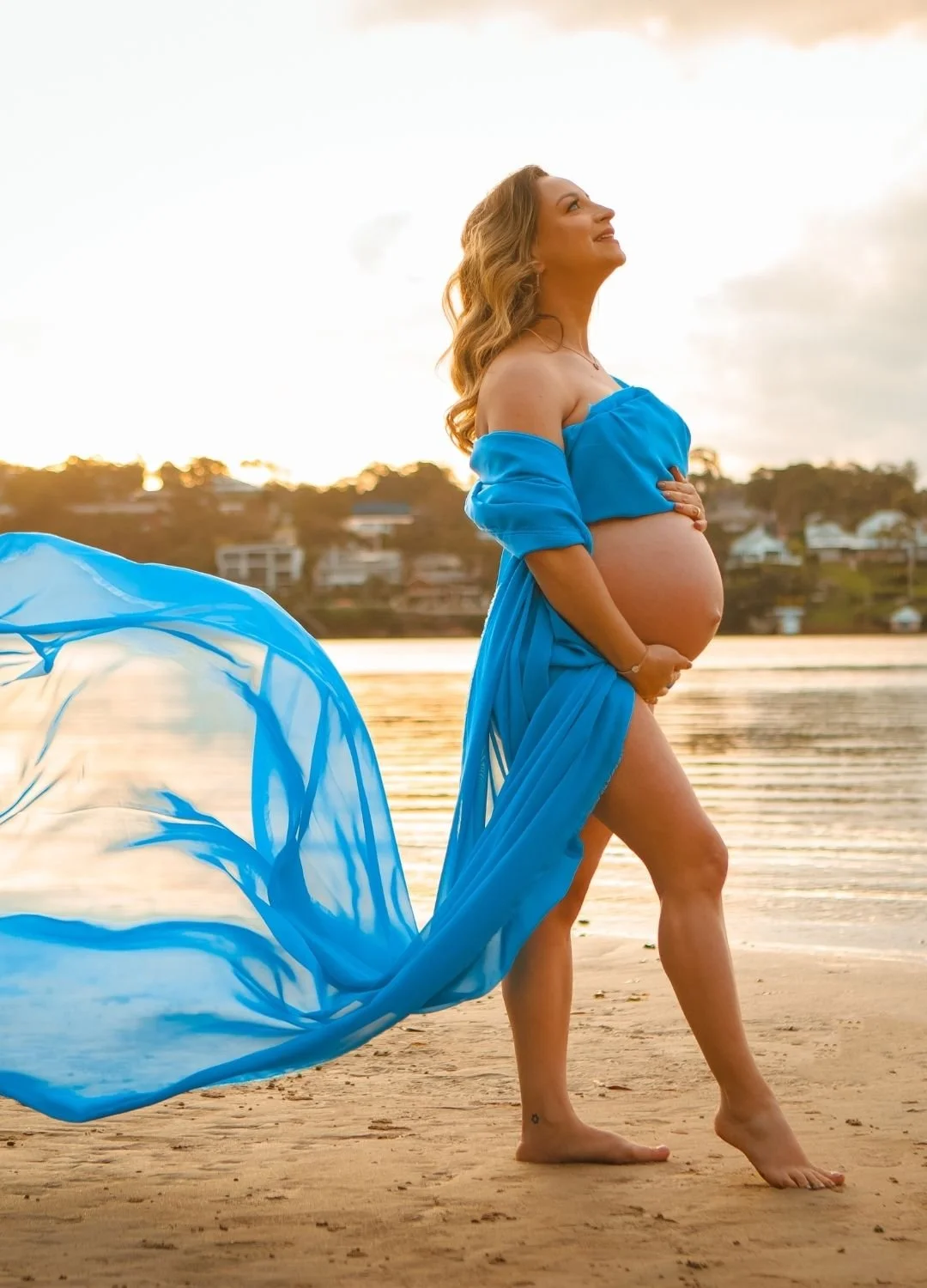 Pregnant woman in a blue dress standing on a beach at sunset, holding her belly and smiling, with flowing fabric and water in the background.
