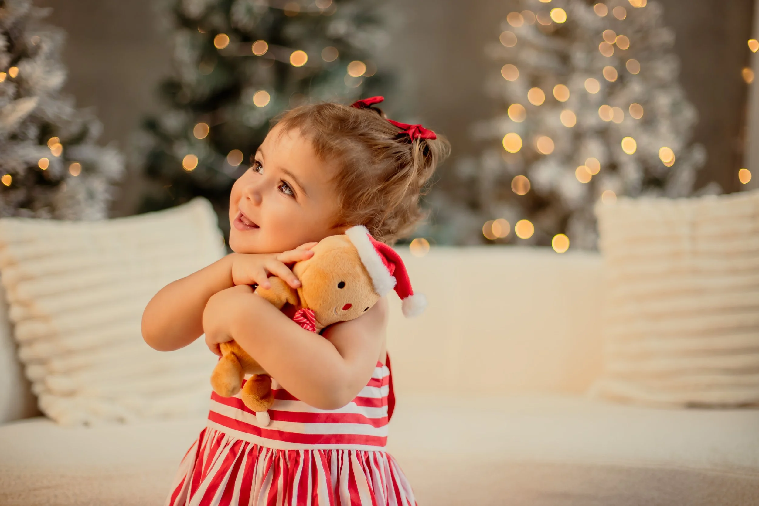 A young girl with curly hair wearing a red and white striped dress, holding a stuffed reindeer toy with a Santa hat, sitting on a beige couch in front of decorated Christmas trees with holiday lights.