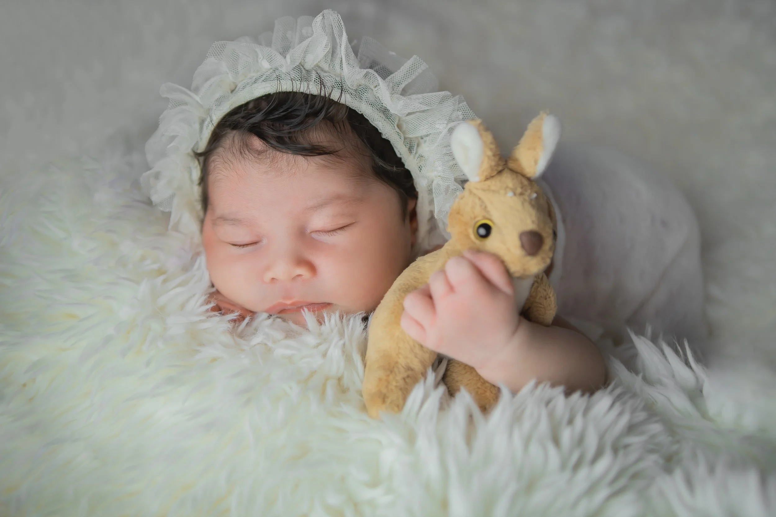 A sleeping baby wearing a ruffled bonnet, lying on a soft, fluffy blanket, holding a small stuffed deer plush toy.