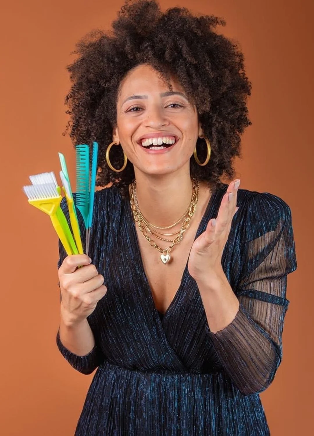 A woman with curly hair, wearing gold hoop earrings, layered gold necklaces, and a dark blue dress, holds colorful hair coloring tools and smiles brightly against a brown background.