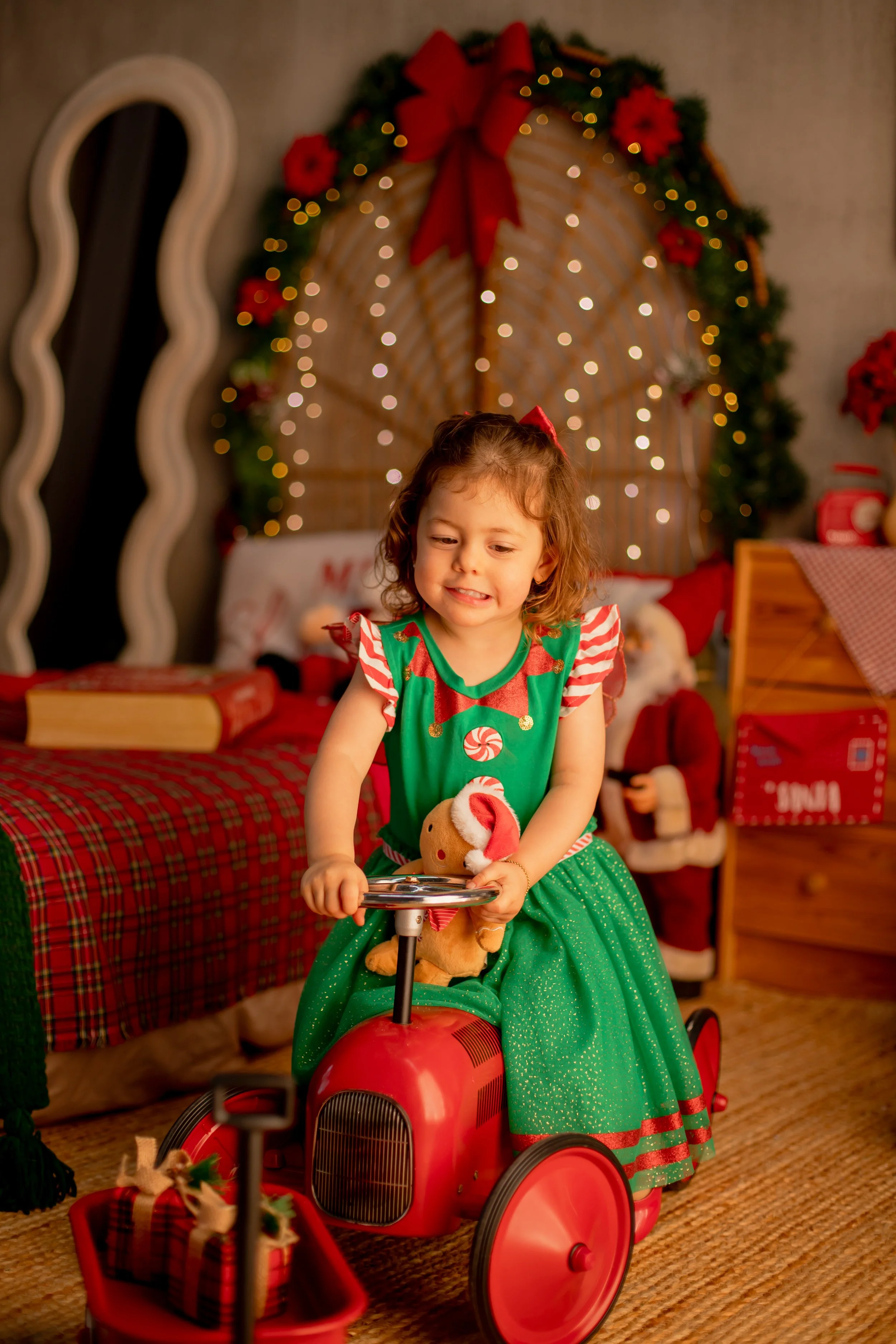 A young girl in a festive elf costume riding a red tricycle with a plush Christmas toy, surrounded by Christmas decorations including a wreath with a red bow and lights, and holiday-themed gifts.