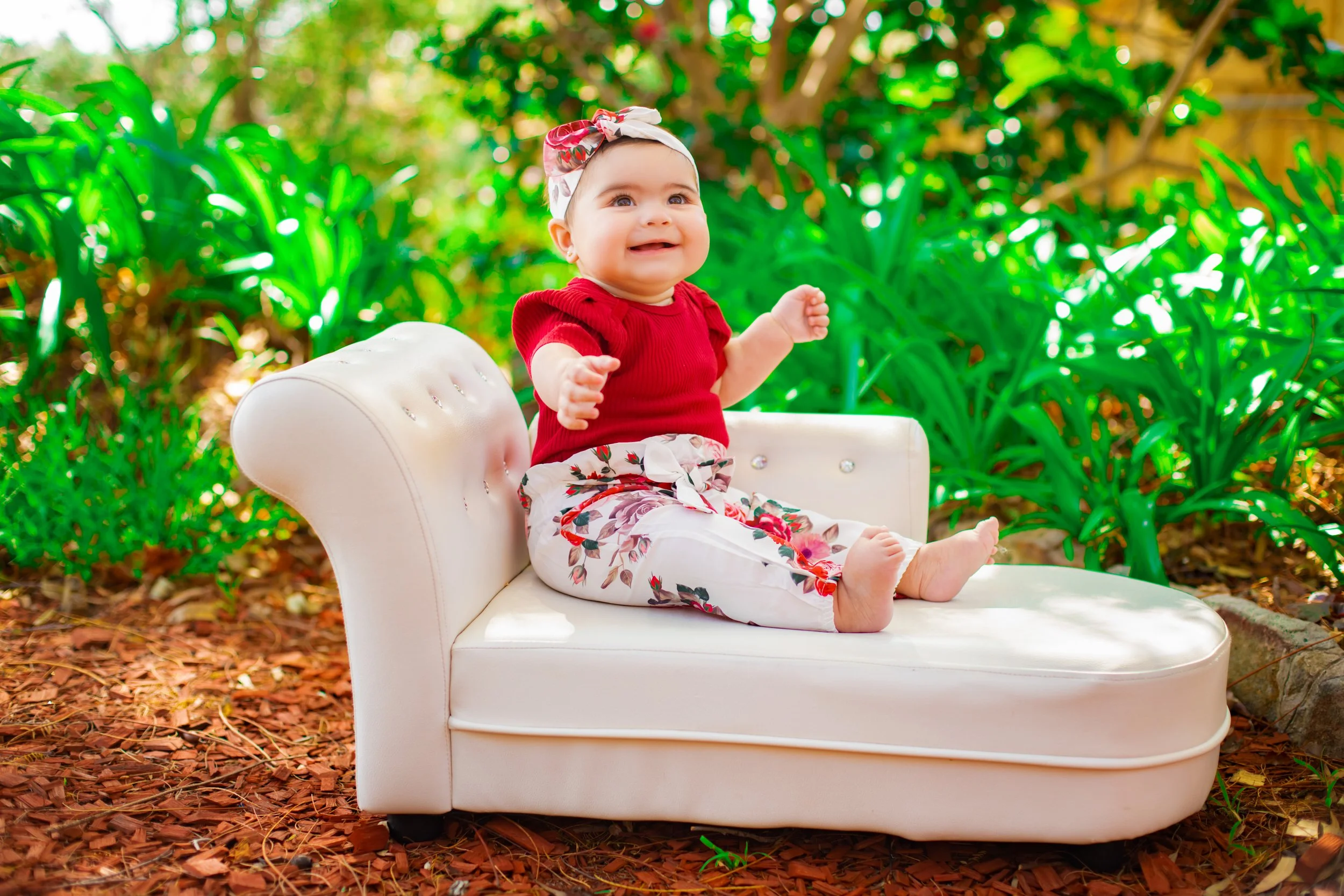 A baby girl sitting on a white sofa outdoors surrounded by green plants, wearing a red top, floral pants, and a white headband, smiling happily.