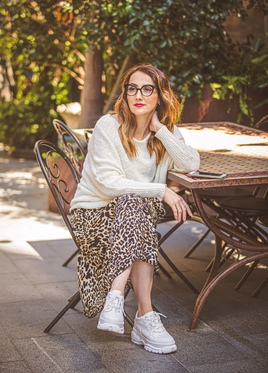 Woman with glasses wearing a white sweater and leopard-print skirt, sitting outdoors at a table with trees and foliage in the background.