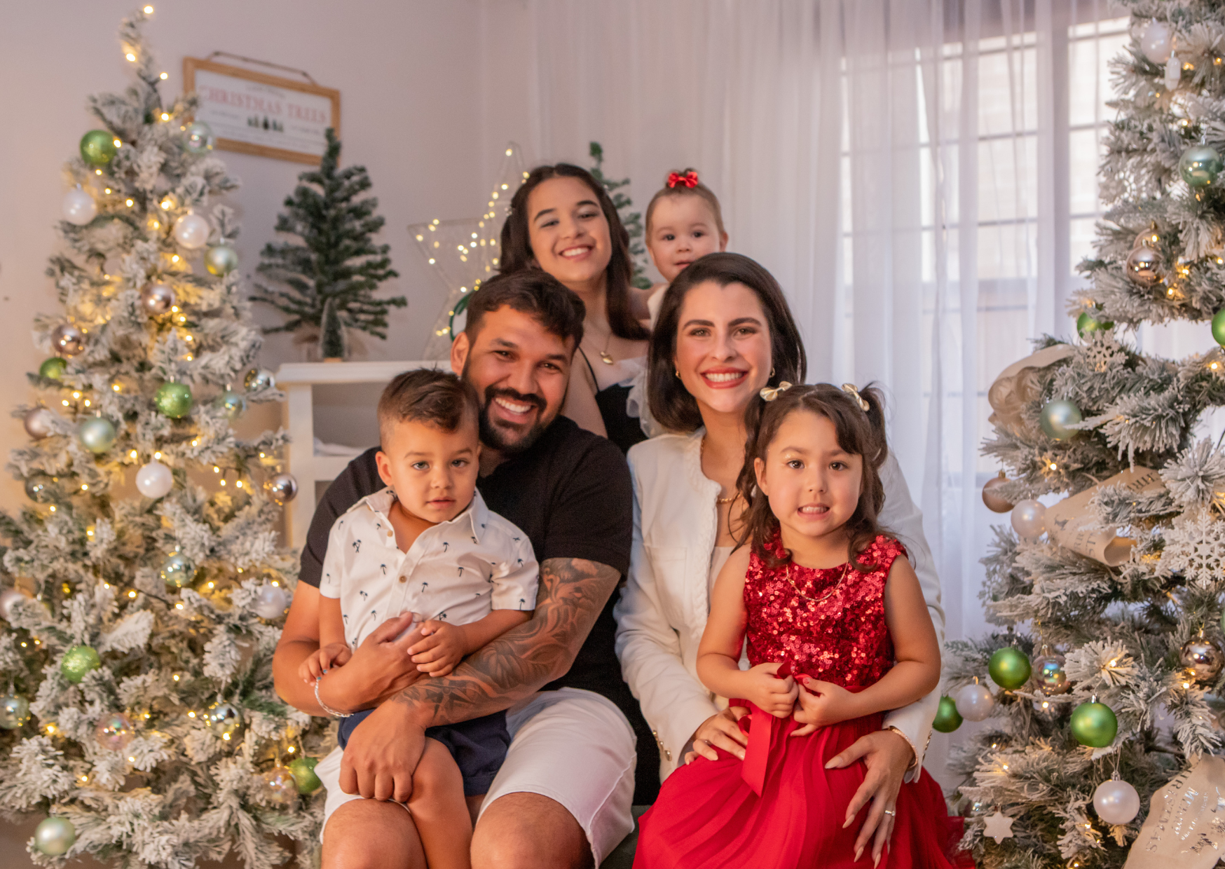 A smiling family of seven posing together in a festive Christmas setting with decorated Christmas trees and white curtains in the background.