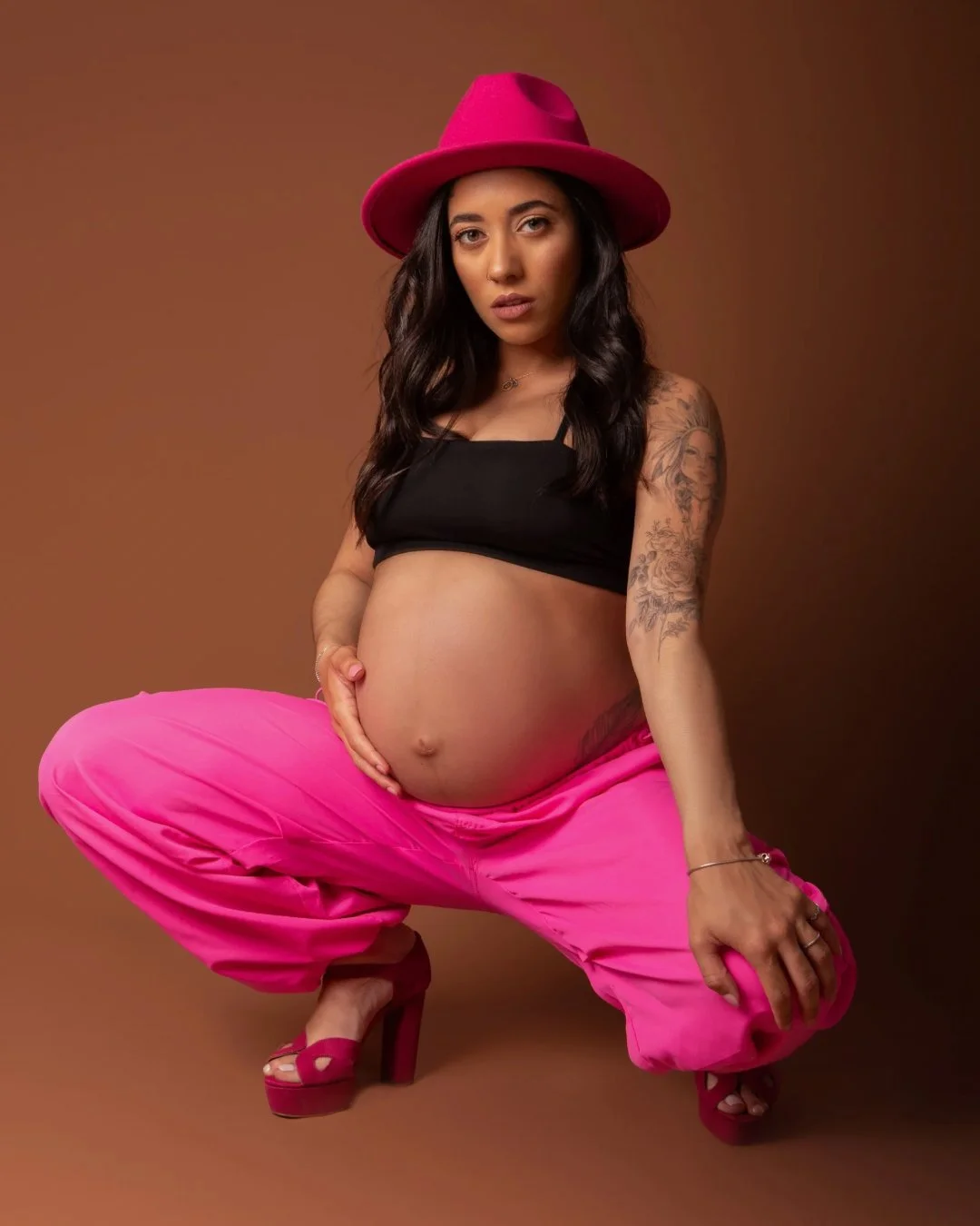 Pregnant woman with long dark hair, wearing a pink hat, black top, pink pants, and high heels, posing against a brown background.