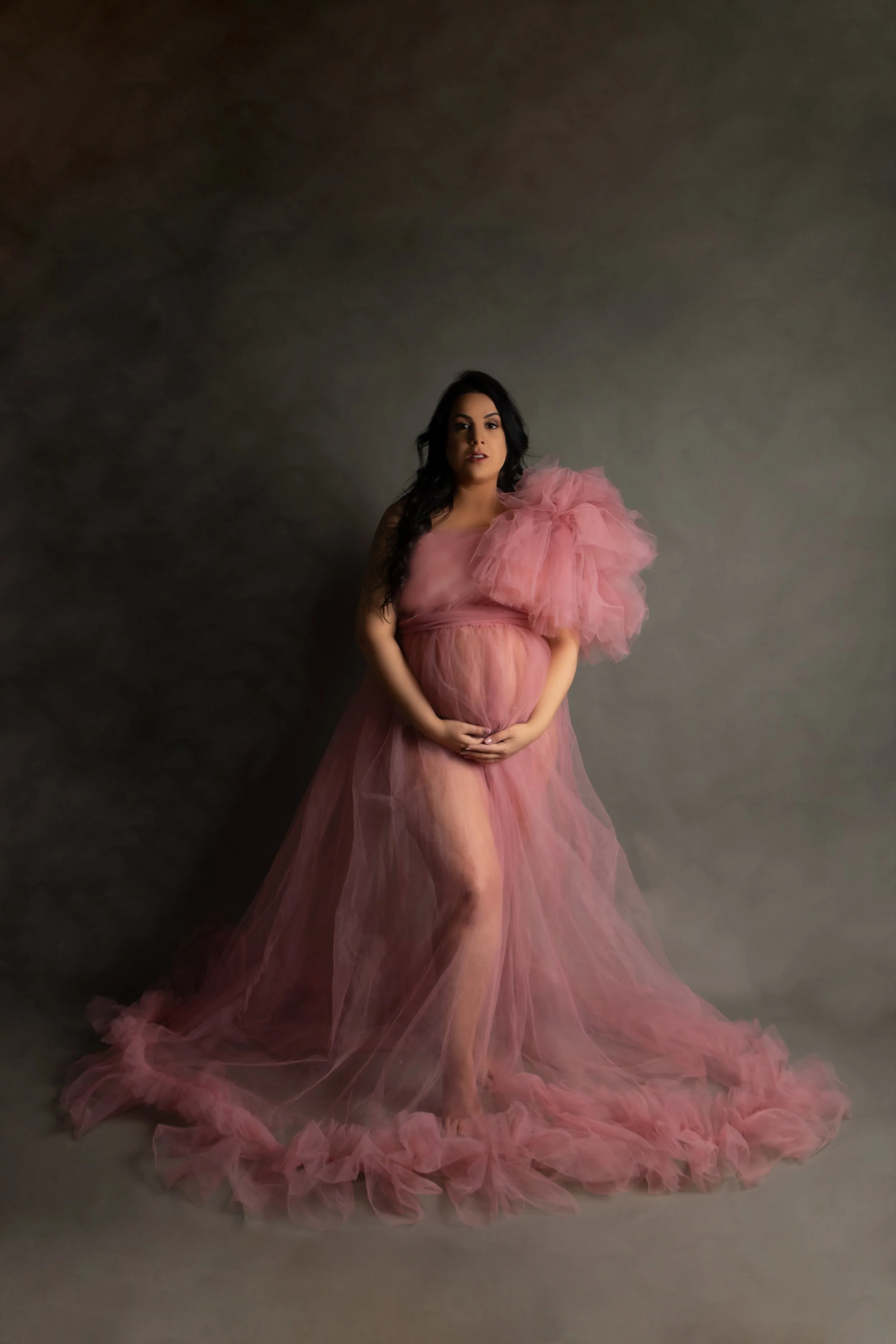 A pregnant woman wearing a flowing, pink tulle gown with a large ruffle on her shoulder, standing against a plain grey background.