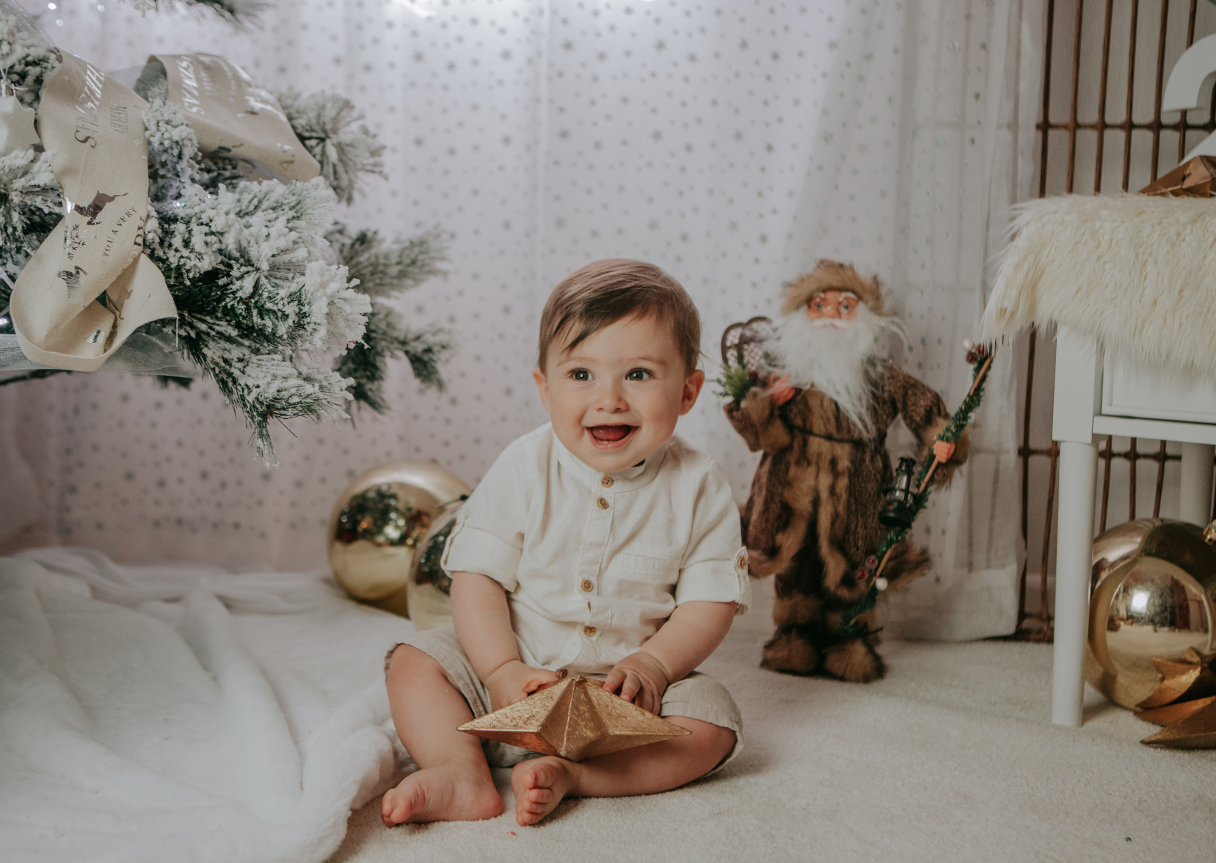 A smiling young boy is sitting on the floor in front of a decorated Christmas tree, holding a gold gift box, with a Santa Claus decoration and gold ornaments in the background.