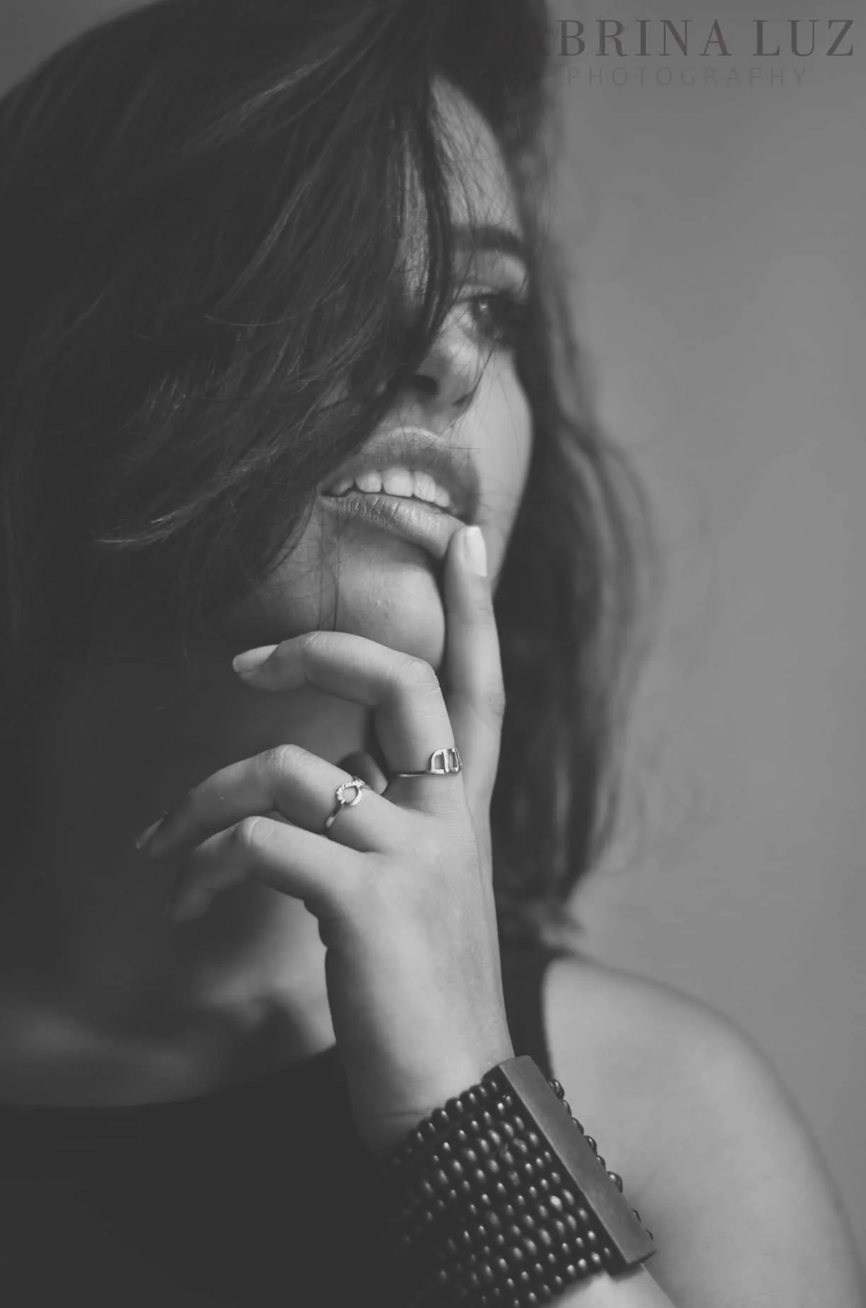 Black and white photograph of a woman with wavy hair, holding her finger to her lips, wearing rings and a beaded bracelet.