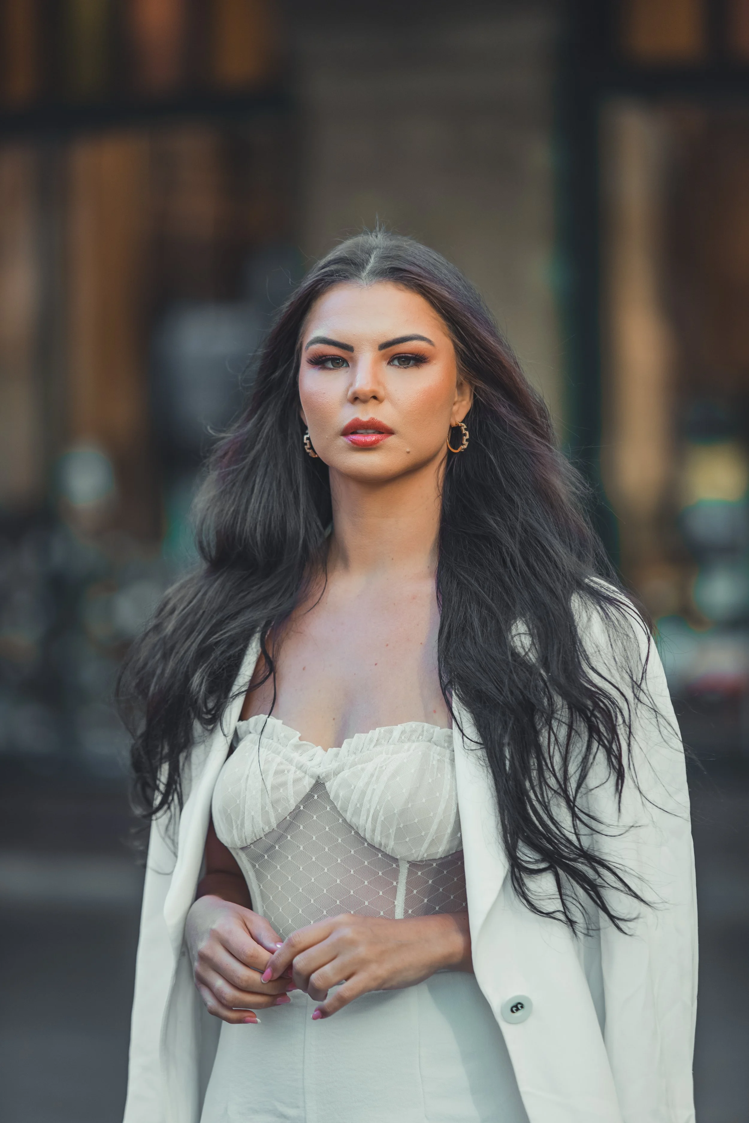 A woman with long dark hair, wearing a beige top and white blazer, standing outdoors in an urban setting.