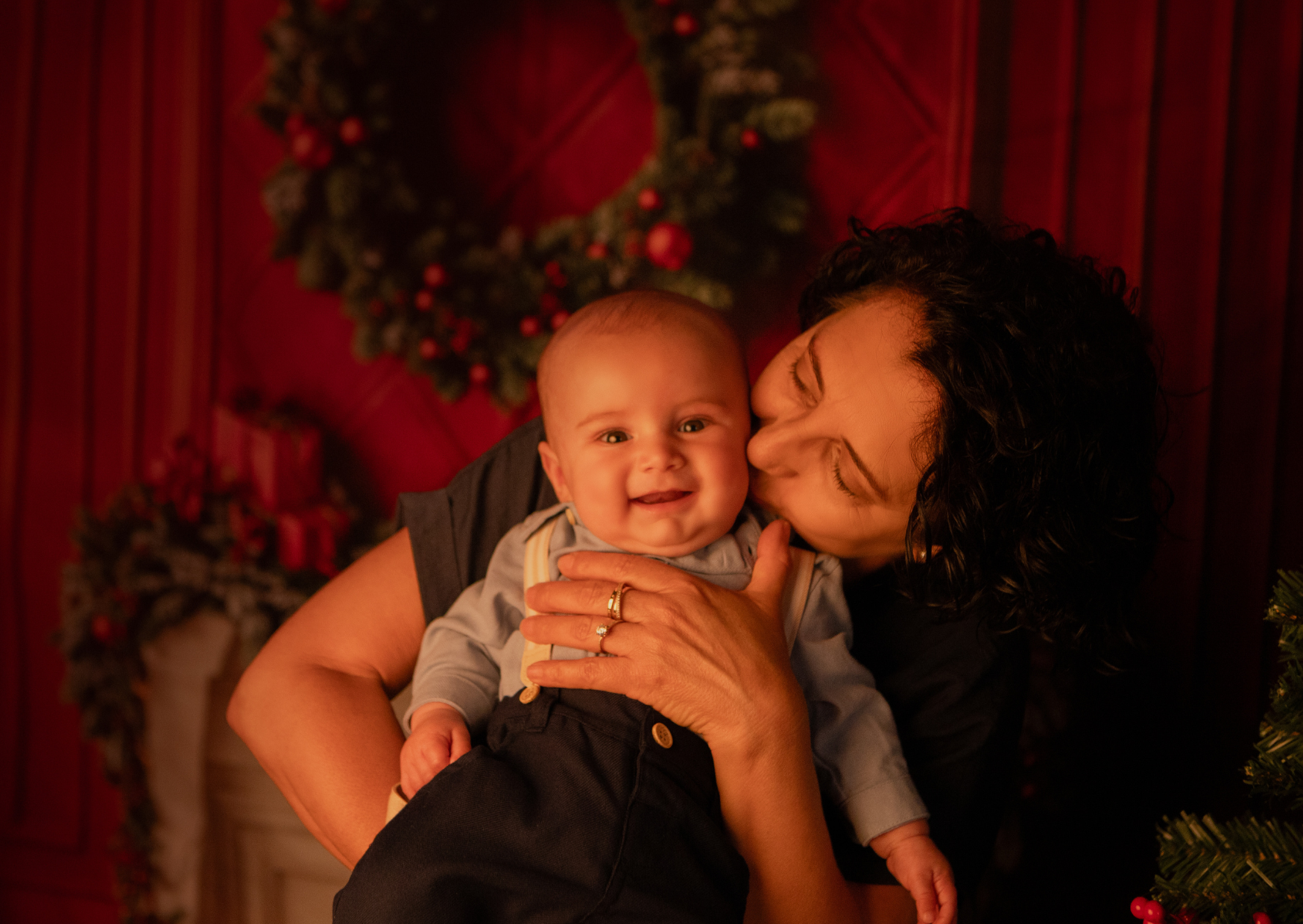 A woman kissing a smiling baby boy on the cheek inside a room decorated for Christmas with a wreath and Christmas tree.