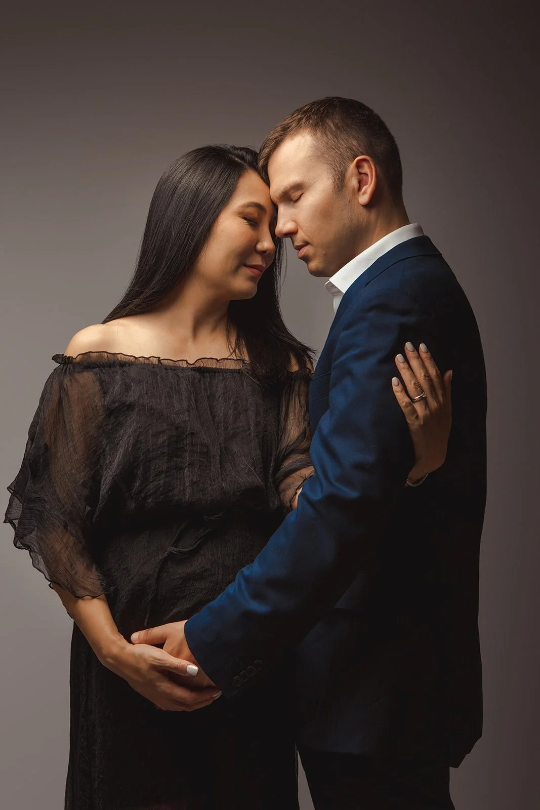 A couple with their foreheads touching, eyes closed, holding hands in a romantic pose. The woman has long dark hair and is wearing a black off-shoulder dress. The man has short brown hair and is wearing a navy blue blazer and white shirt.