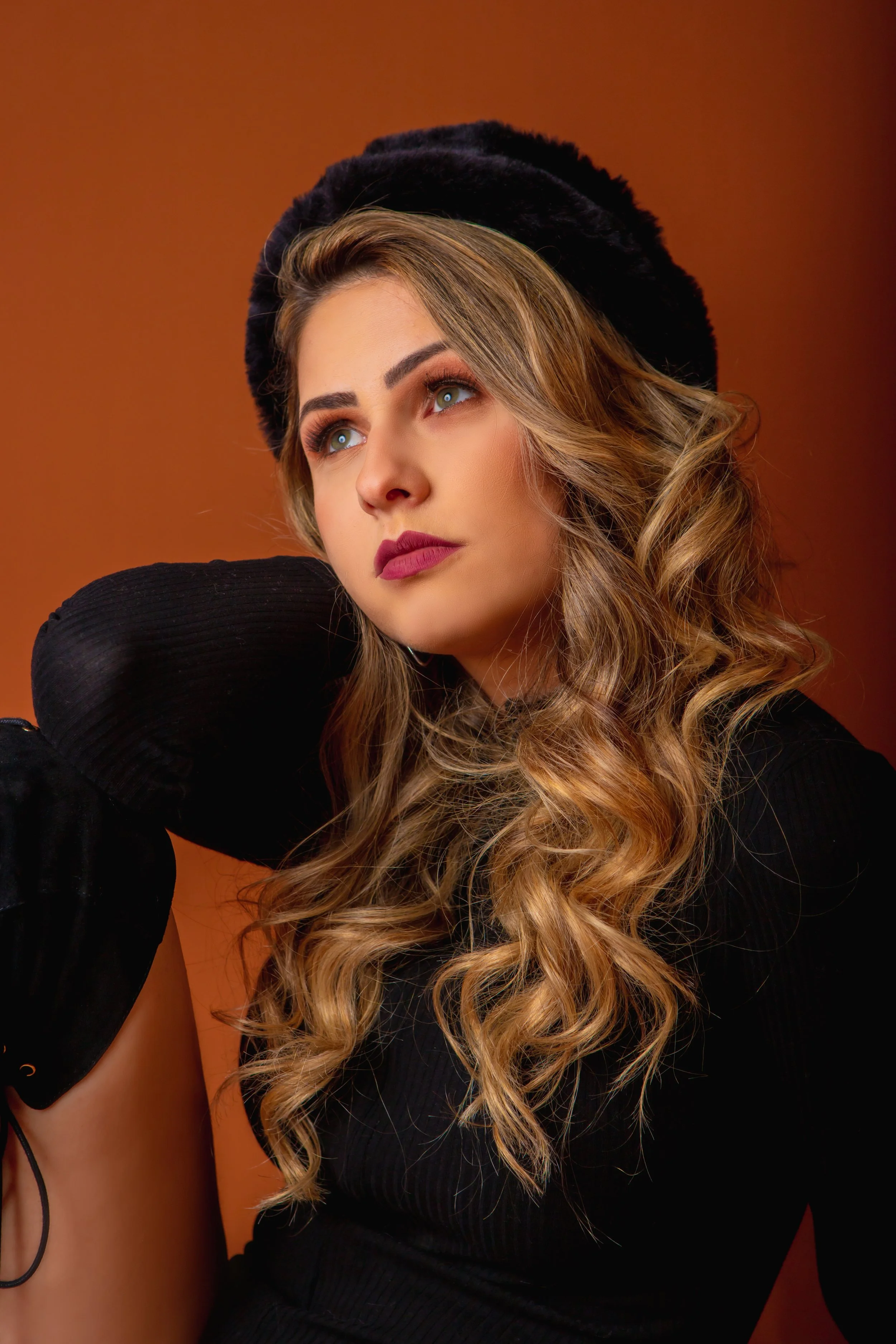 A woman with long wavy blonde hair, wearing a black beret and black top, looking upward with her hand resting on her neck, in front of an orange-brown background.