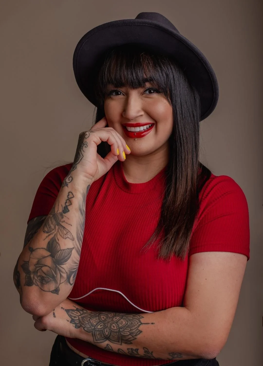 A smiling woman with dark hair, red lipstick, and tattoos on her arms, wearing a black hat and a red shirt, posing with her hand near her face against a plain background.