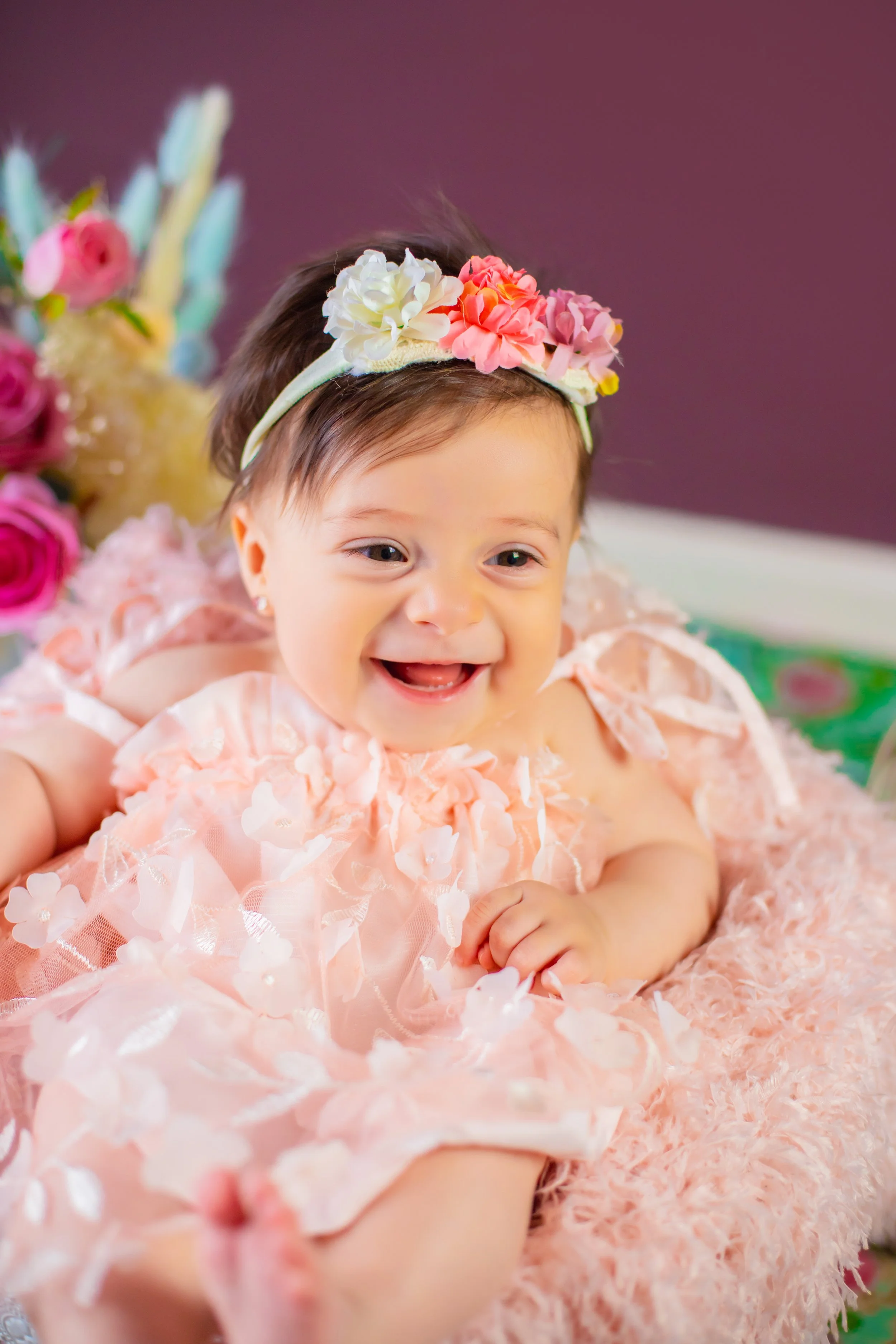 A smiling baby girl wearing a pink floral dress and a flower headband, lying on a pink blanket with birthday decorations and a birthday cake in the background.