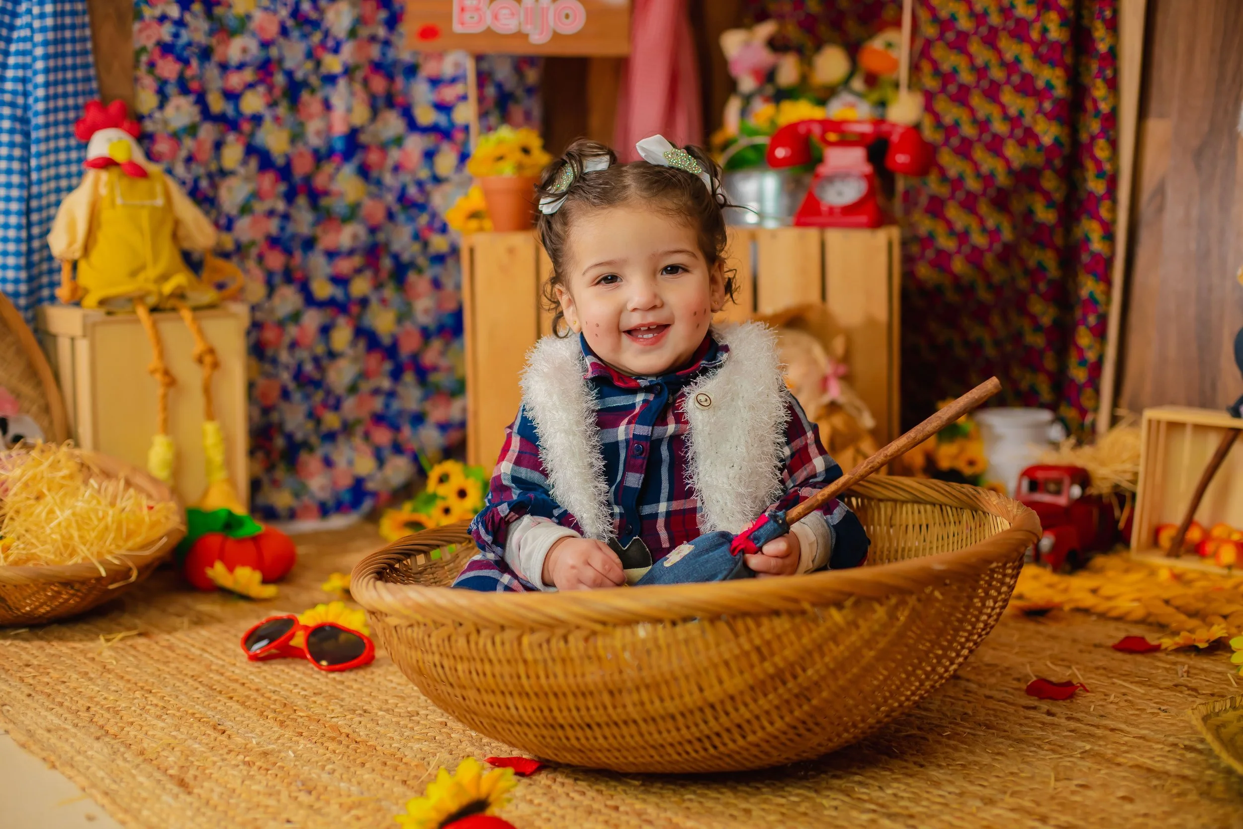 Young girl with dark curly hair, wearing a plaid shirt and fuzzy vest, sitting in a woven basket, smiling in a colorful, autumn-themed setting with toys, flowers, and decorative items around her.