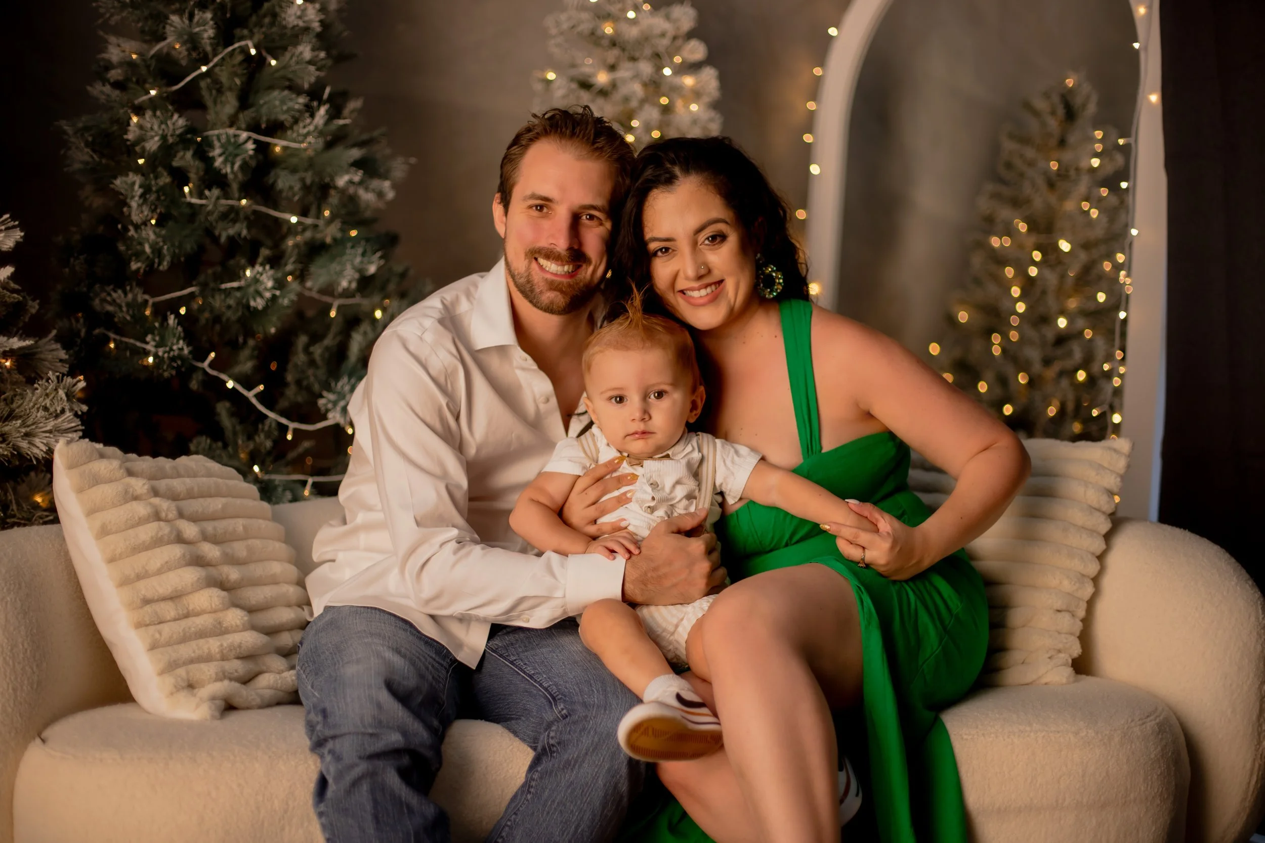 Family sitting on a cream couch with two Christmas trees and fairy lights in the background, celebrating the holidays.