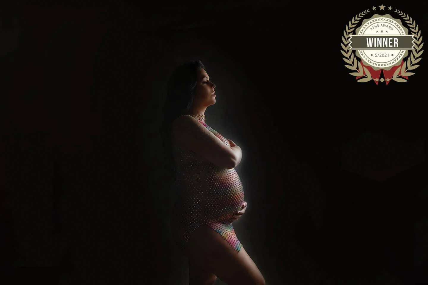 A pregnant woman standing in side profile with arms crossed over her chest, wearing a colorful, see-through dress, illuminated by soft light against a dark background, with a