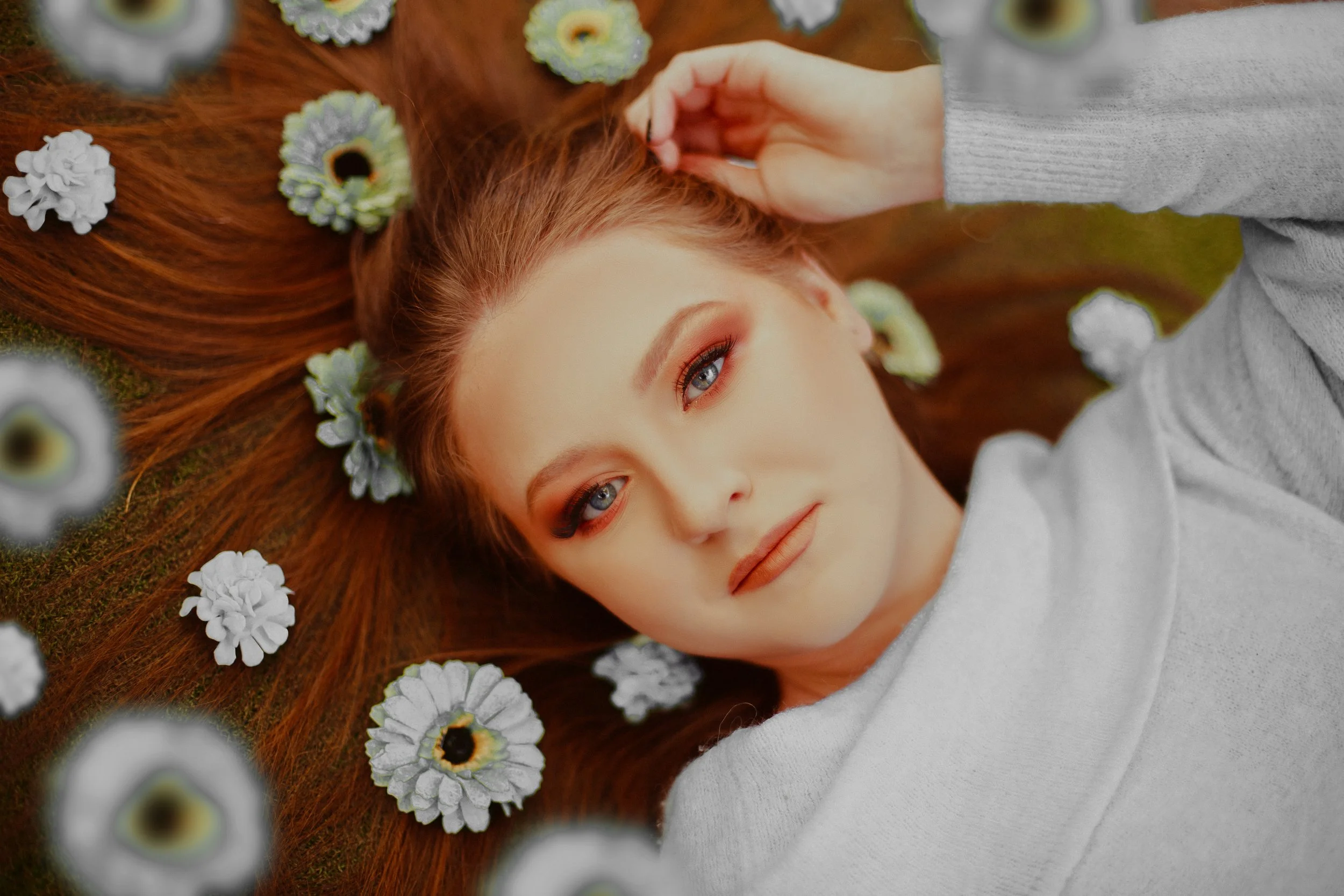 A woman with red hair and blue eyes lying on grass, surrounded by white and yellow flowers, wearing a gray sweater.