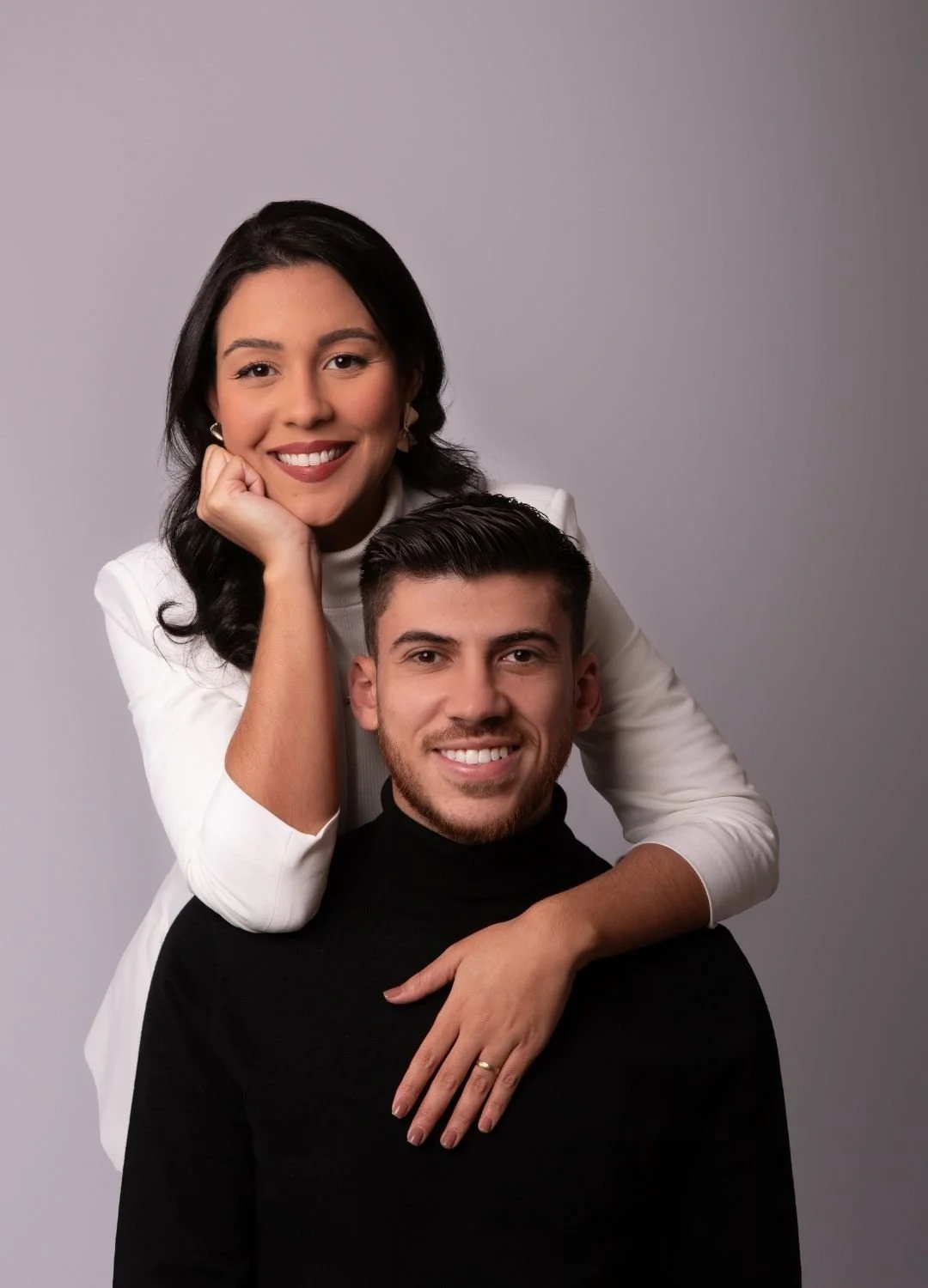 A woman with dark hair and a white blazer leaning on a man's shoulder, who has dark hair and a black turtleneck, both smiling at the camera against a plain gray background.