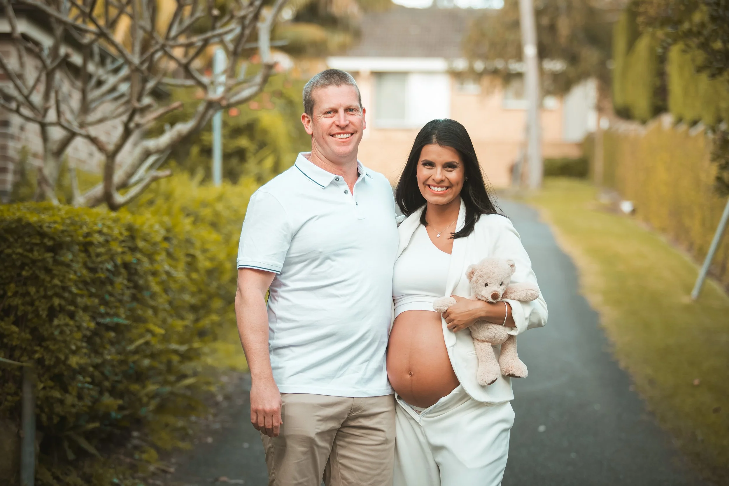 A smiling pregnant woman holding a stuffed teddy bear standing next to a man on a suburban street.