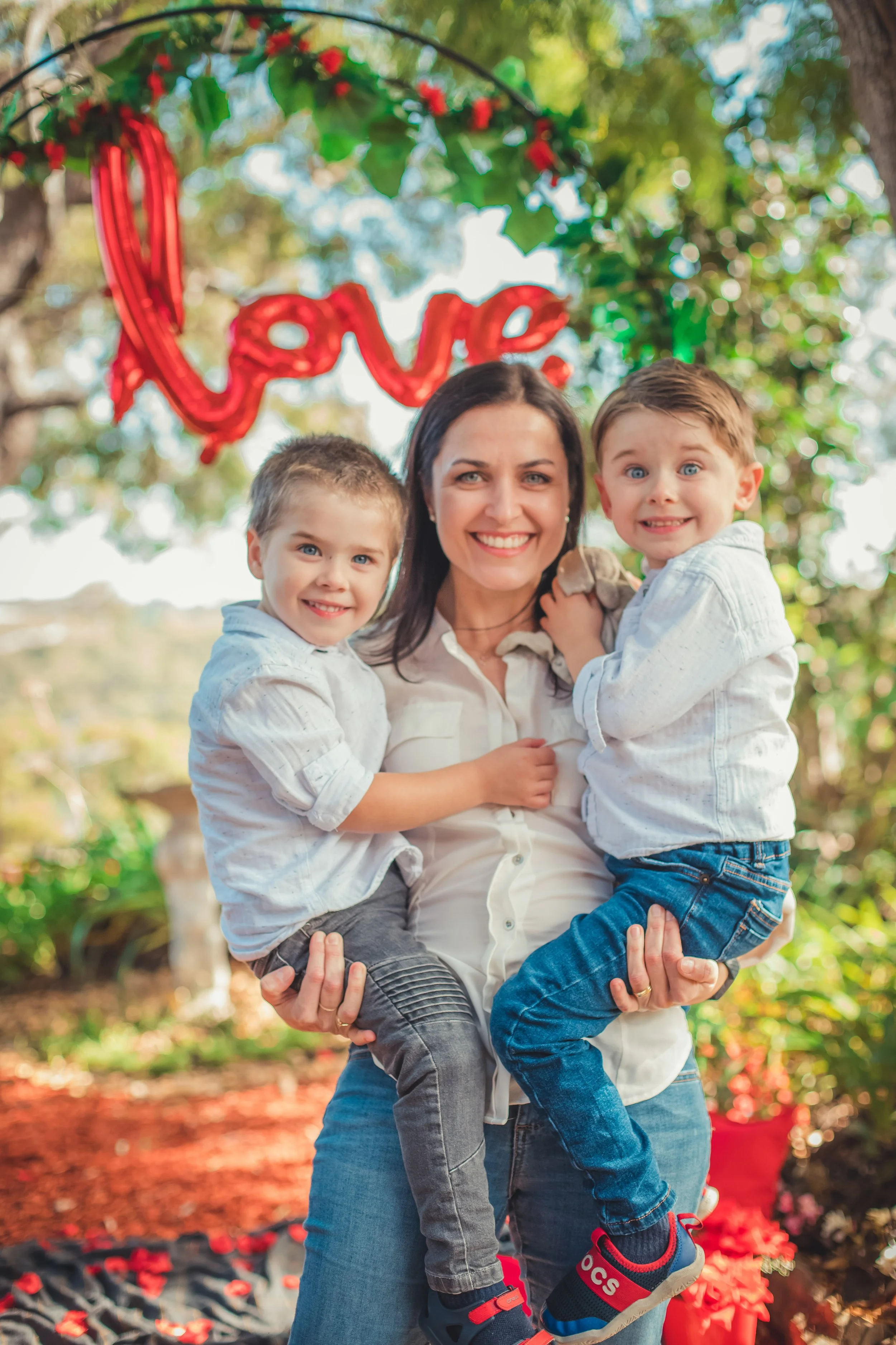 A smiling woman holding two young boys outdoors with a red